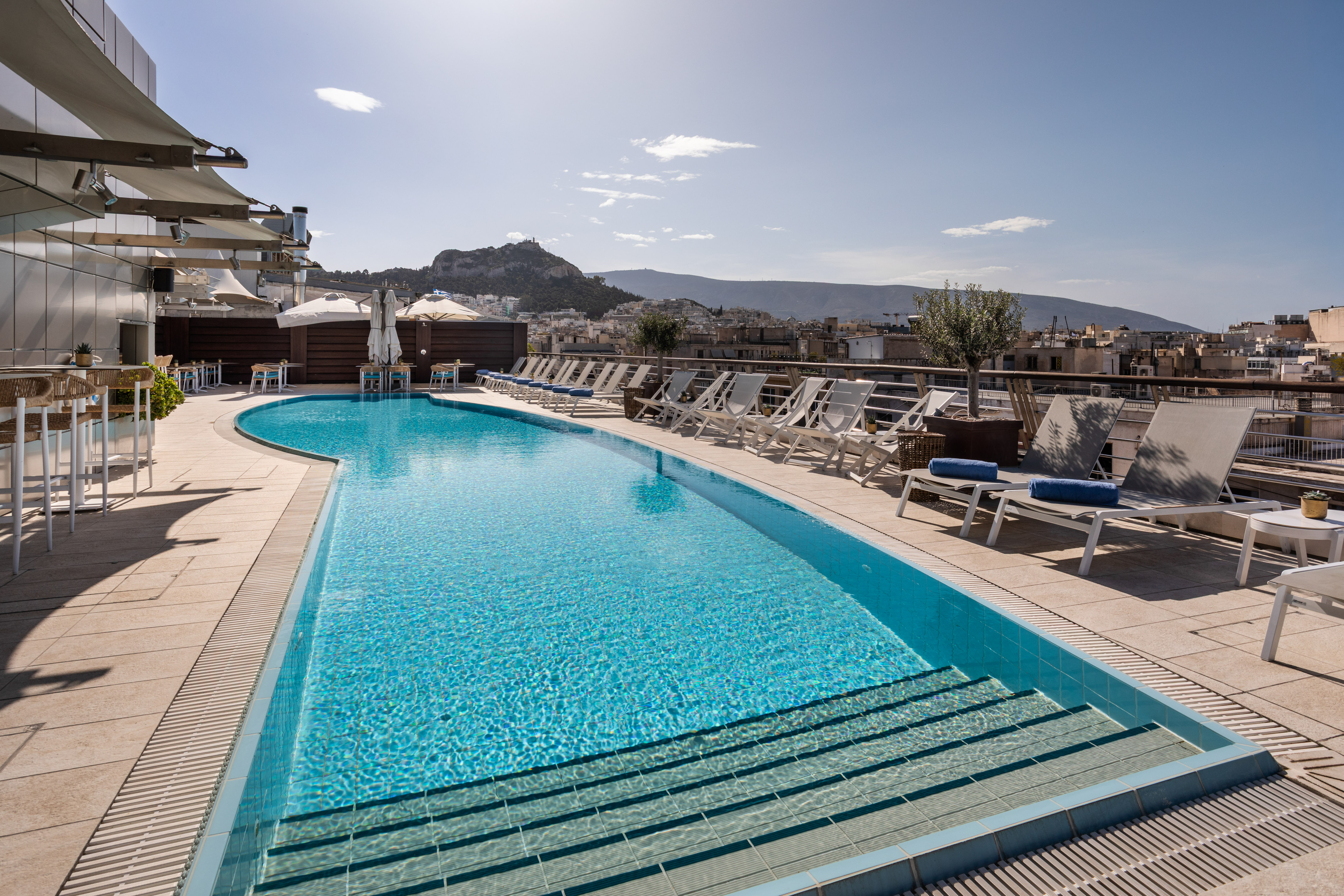 Hotel rooftop pool in Athens with lounge chairs and Acropolis view.