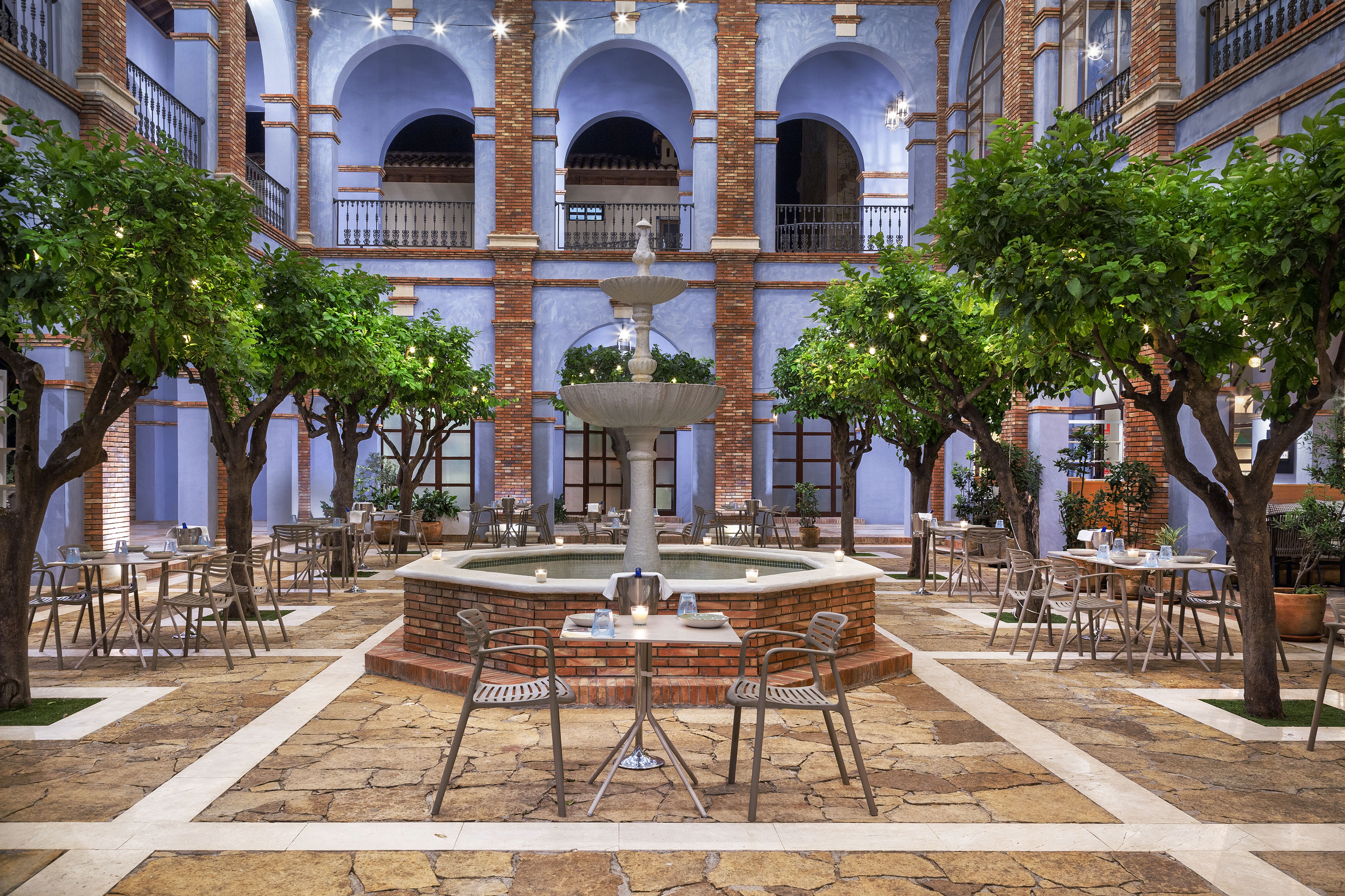Outdoor courtyard dining, central fountain, orange trees, blue arched architecture.