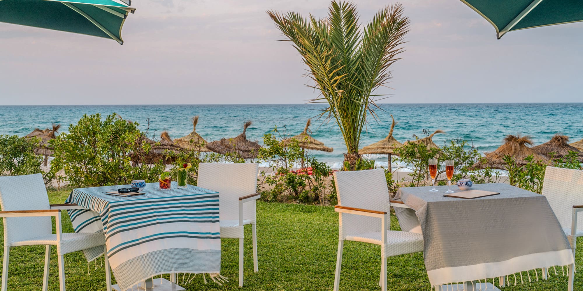 Beachfront outdoor dining tables, ocean view, palm tree, resort umbrellas.