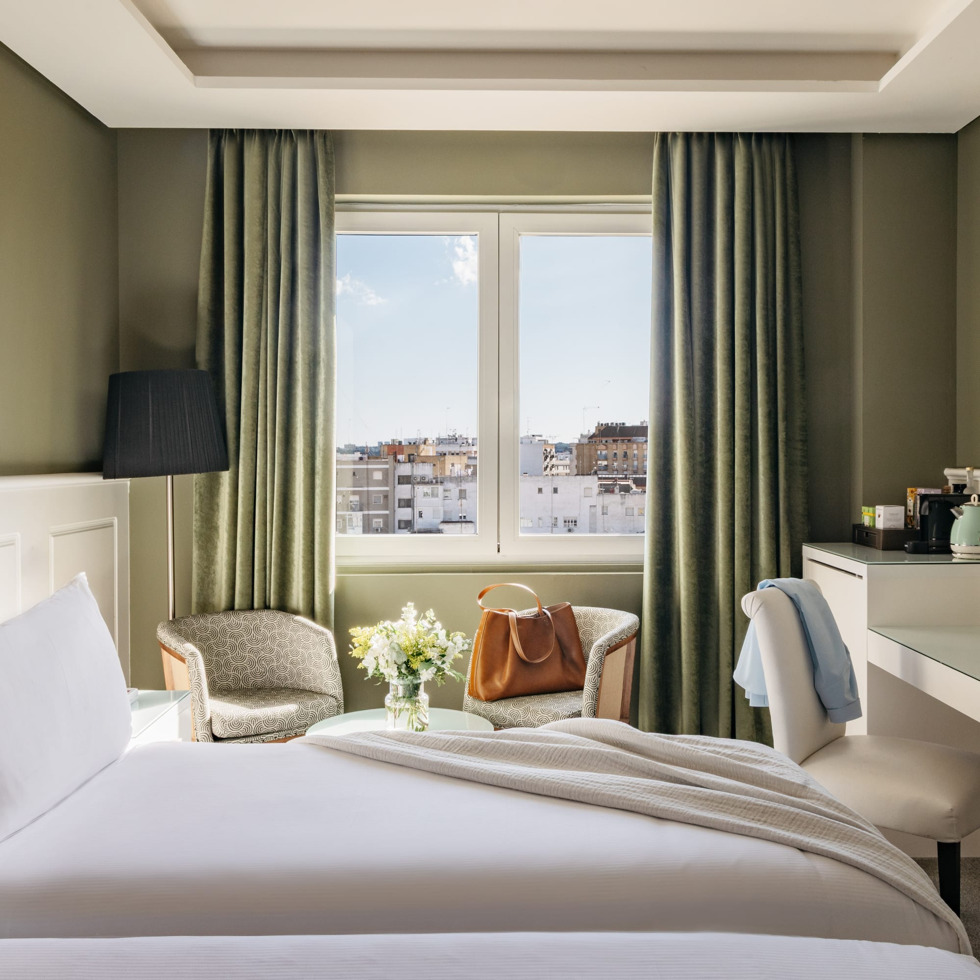 Habitación de hotel con cama, ventana a la ciudad, escritorio y sillones.