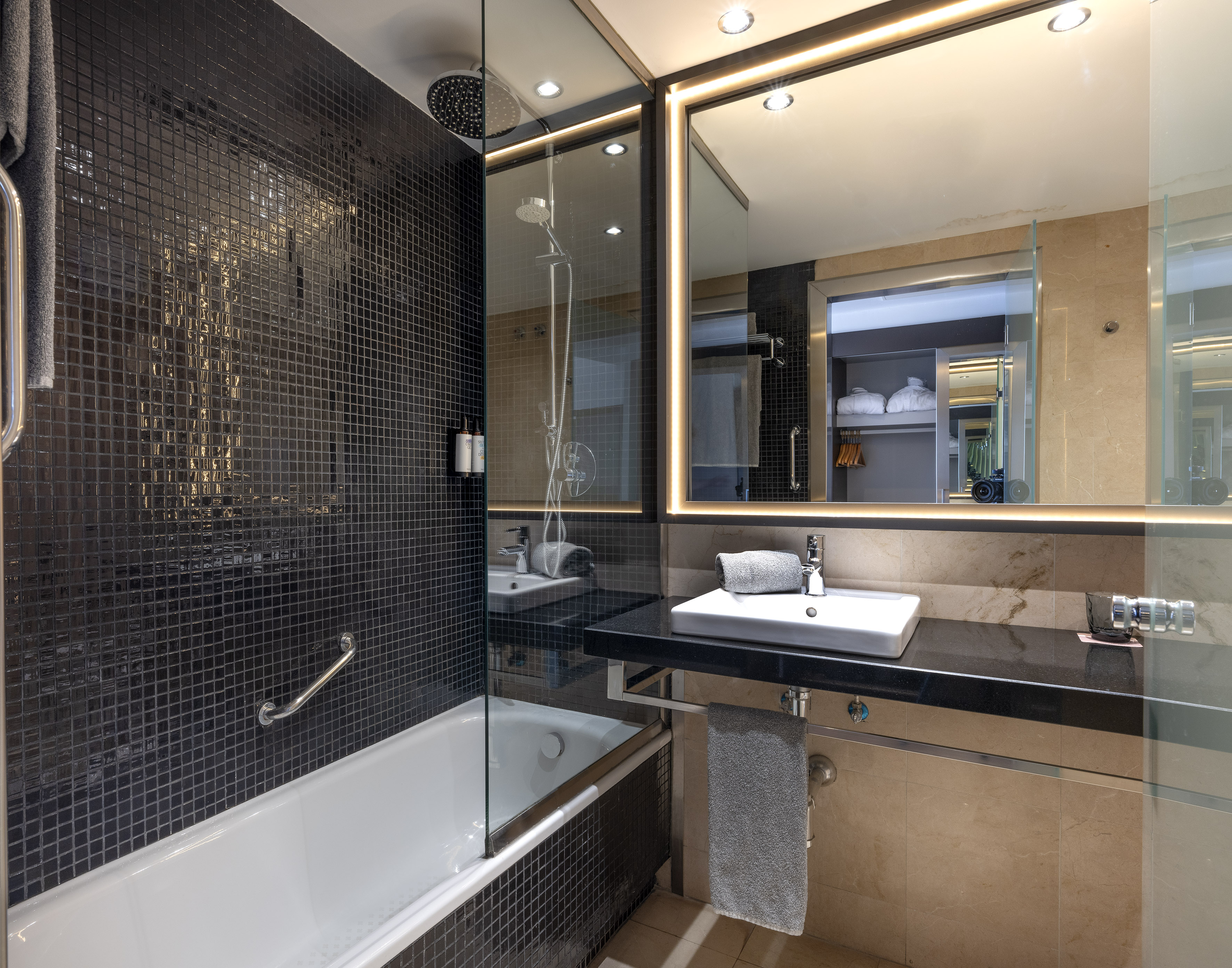 Modern hotel bathroom with black mosaic tiles, white tub, and lighted vanity mirror.