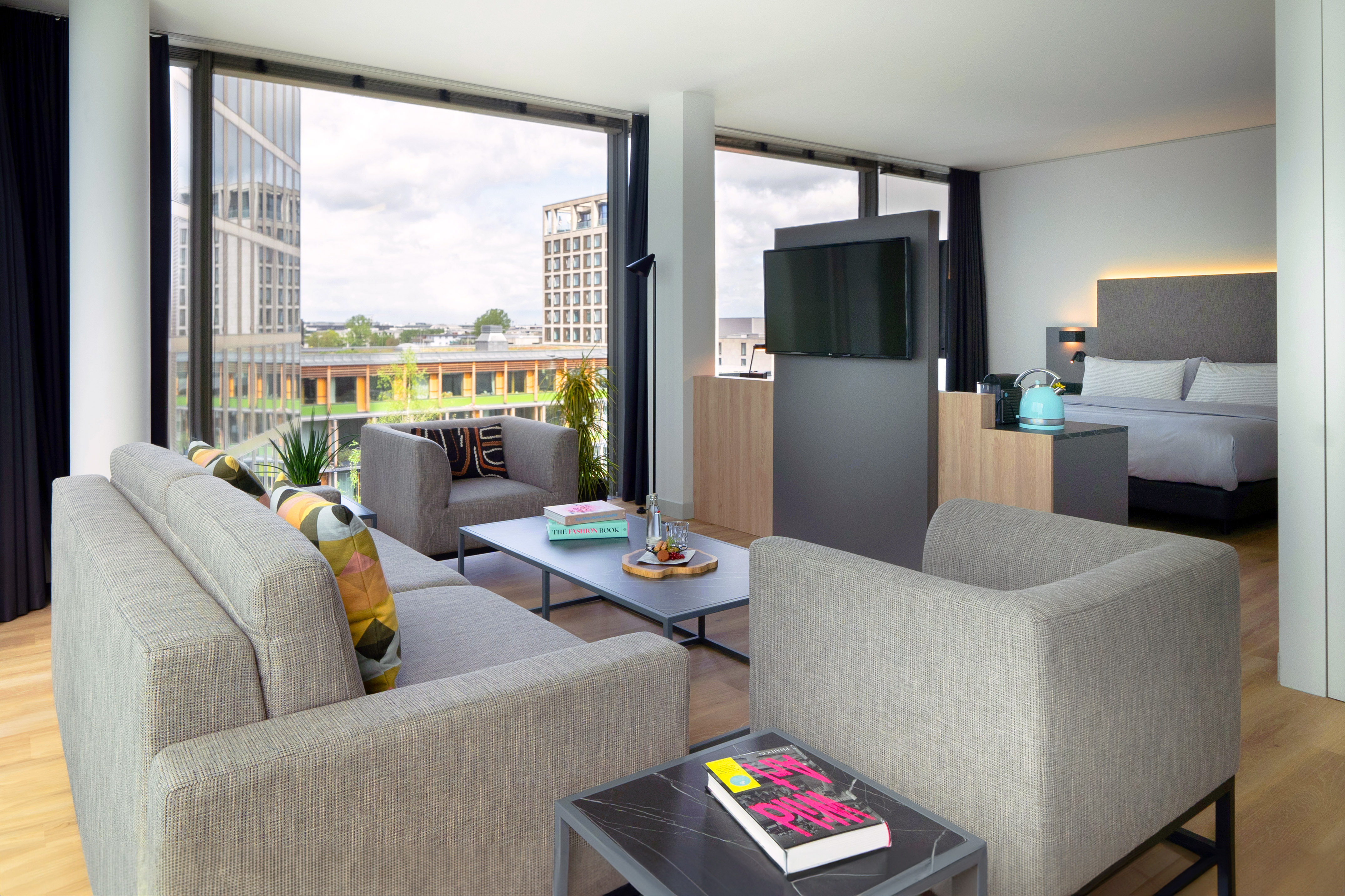 Modern hotel suite: open-plan living room, bedroom, large windows, urban view.