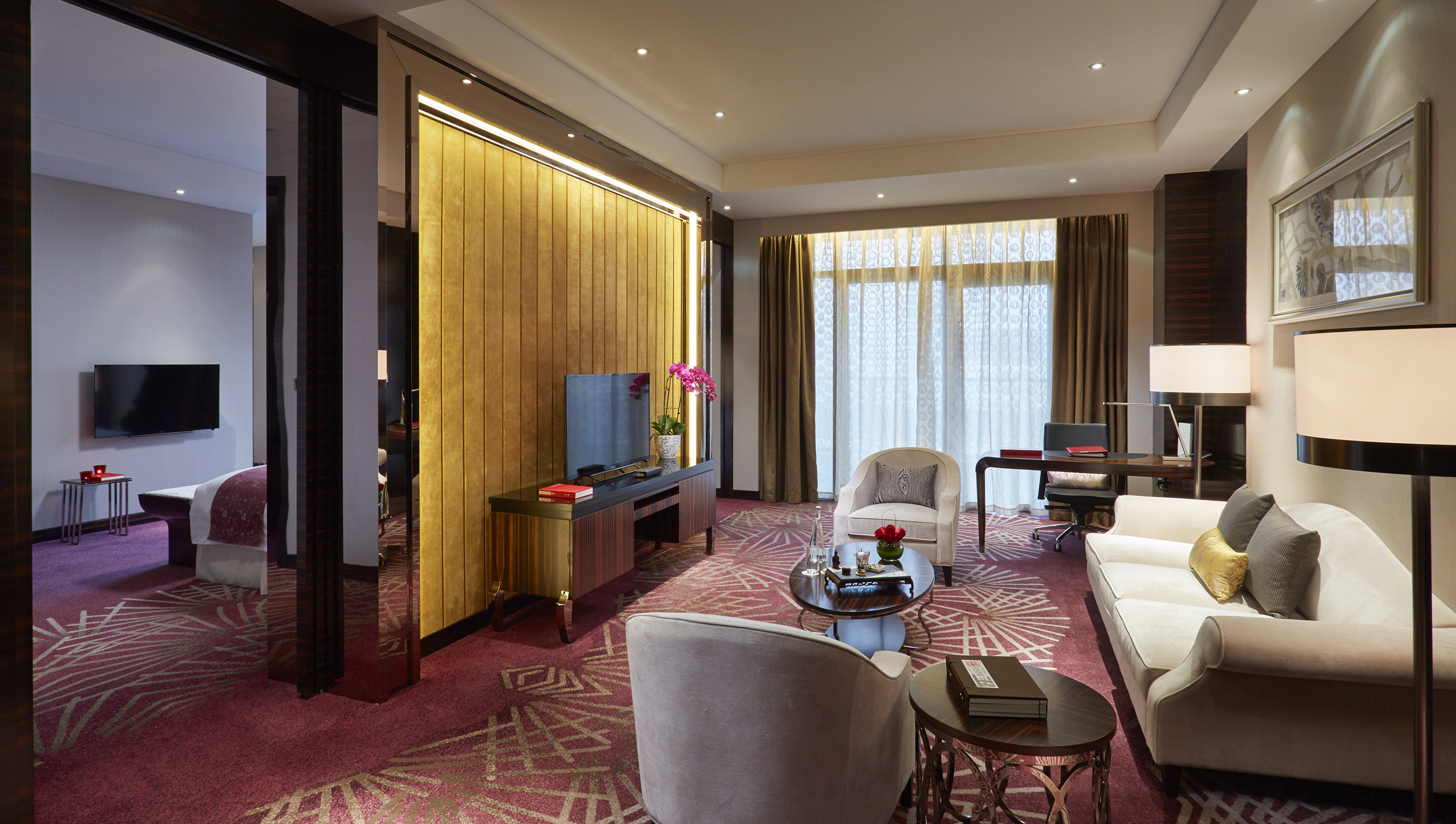 Luxury hotel suite living room with red patterned carpet, sofa, TV, and desk.