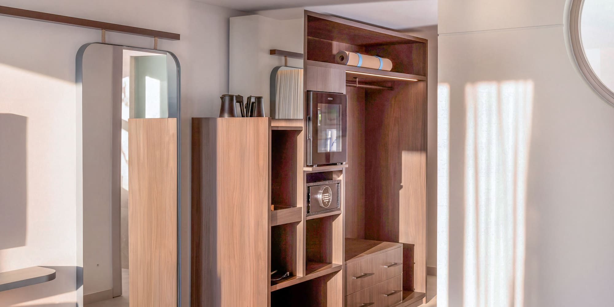 Modern hotel room interior with wooden wardrobe, full-length mirror, minibar, and yoga mat.