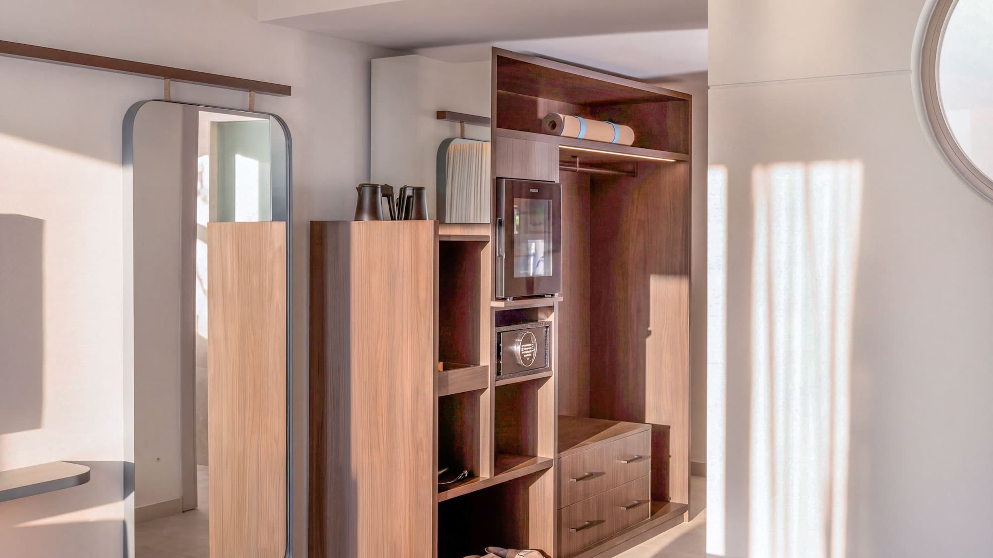 Modern hotel room wardrobe with mirror, minibar, safe, and yoga mat.