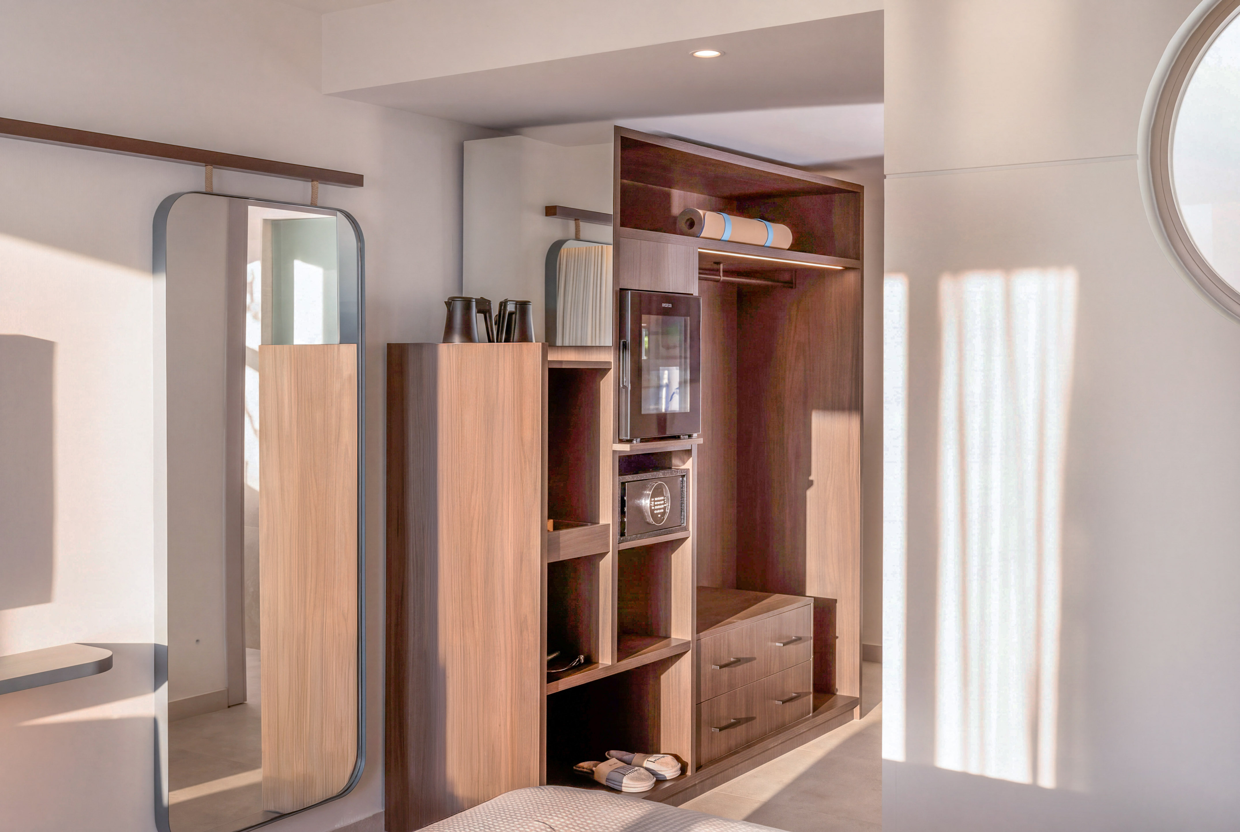 Modern hotel room interior with wooden wardrobe, full-length mirror, minibar, and yoga mat.
