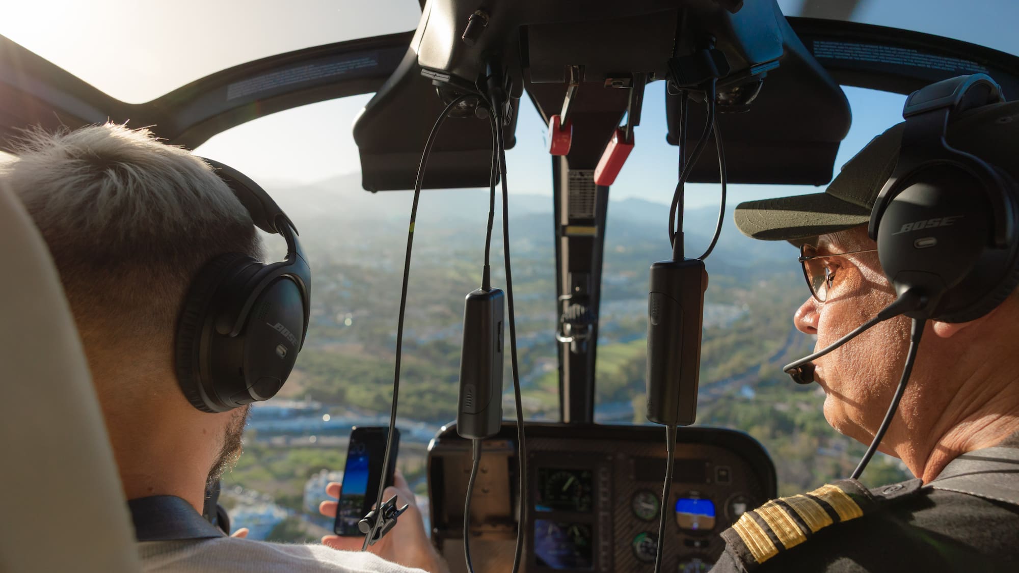 Helicopter pilot & passenger with headsets in cockpit, scenic flight view.