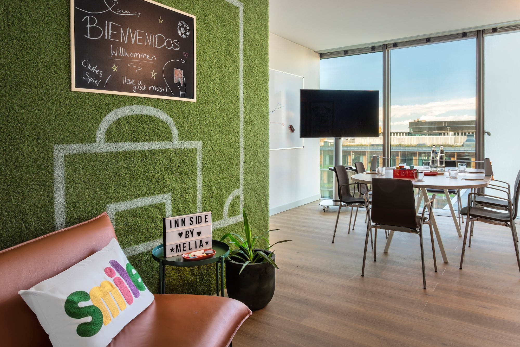 Modern meeting room with a green soccer field wall, welcome sign, and city views.