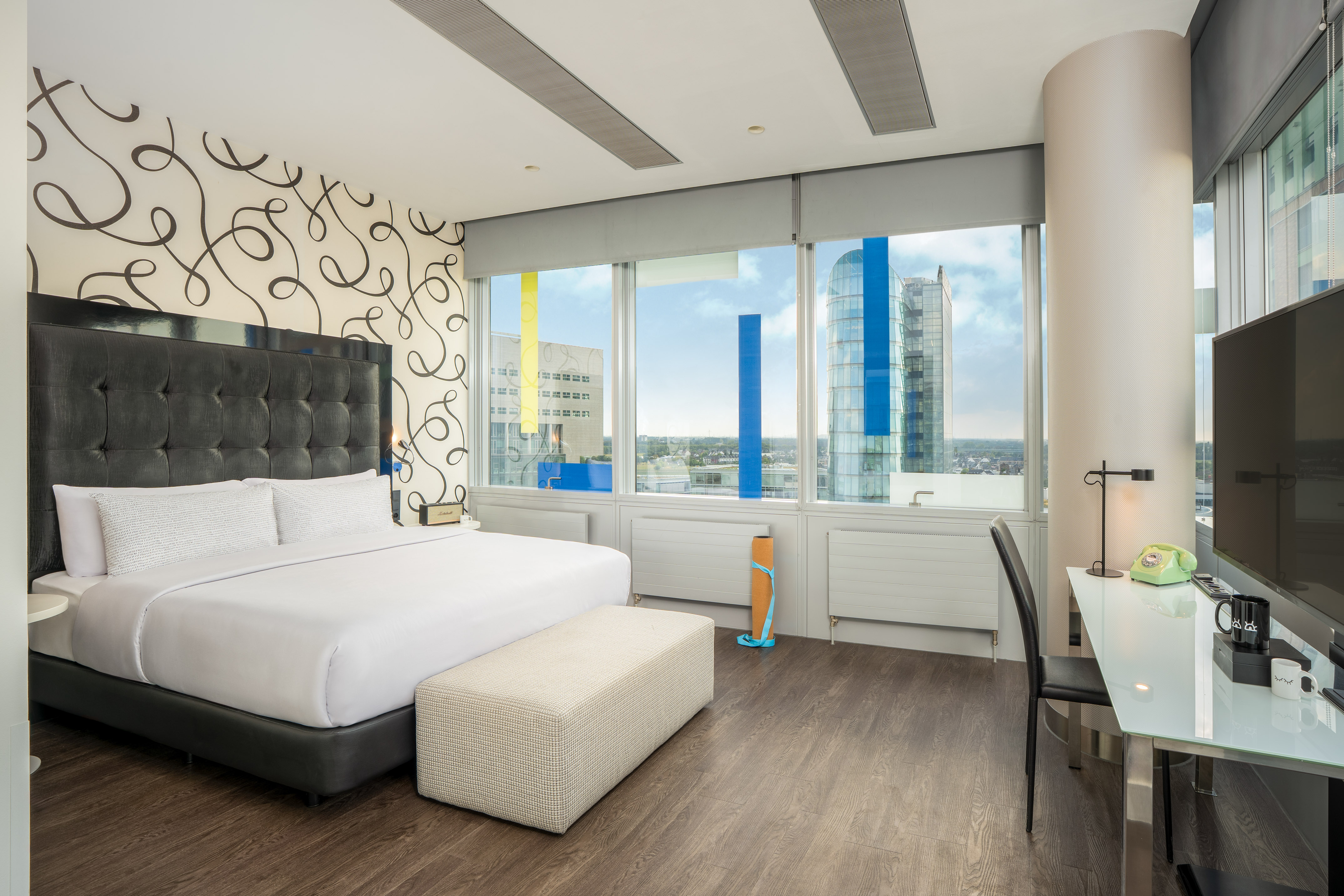 Modern hotel room with king bed, city view, work desk, and patterned wall.