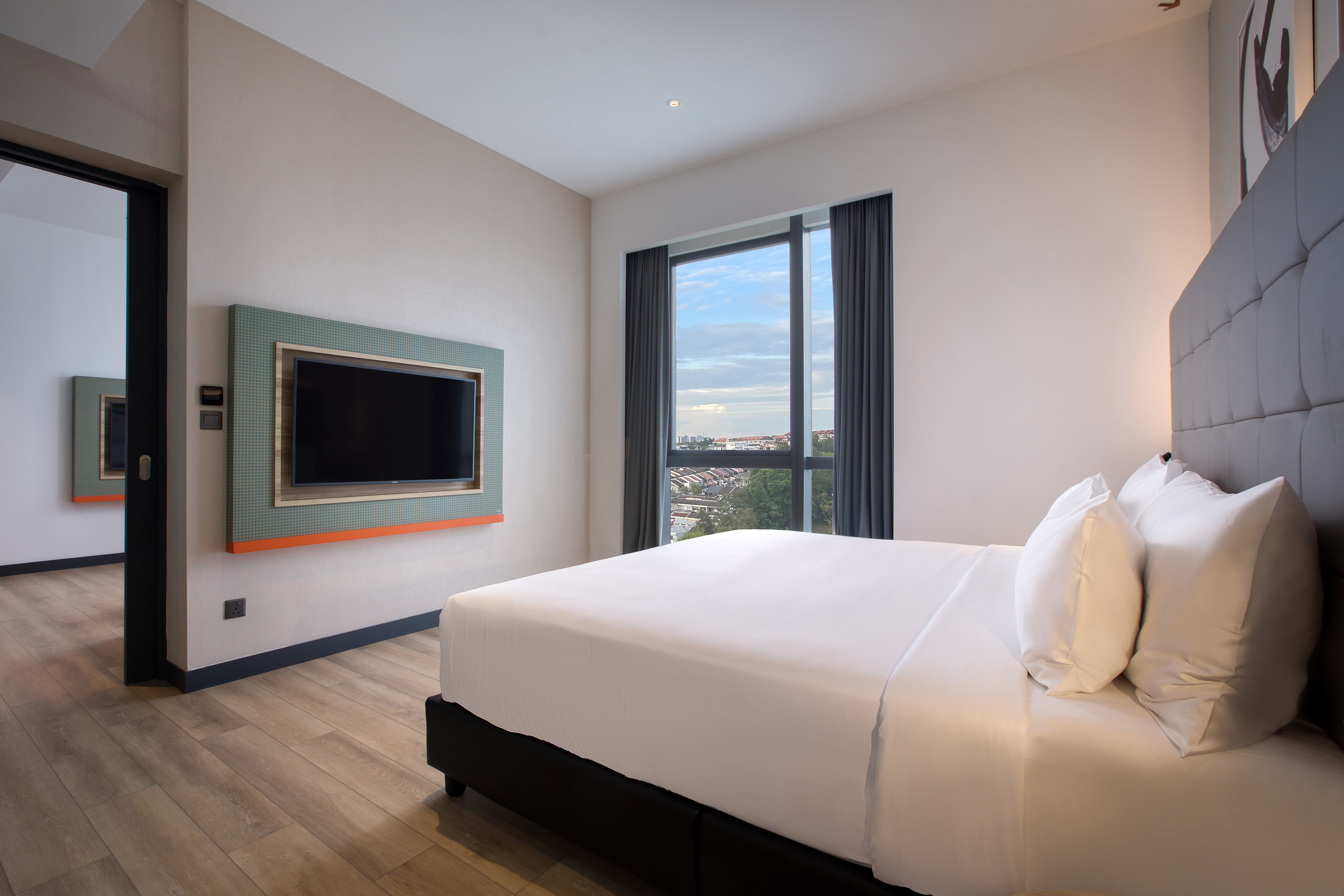 Modern hotel bedroom with white bed, wall-mounted TV, and city view.