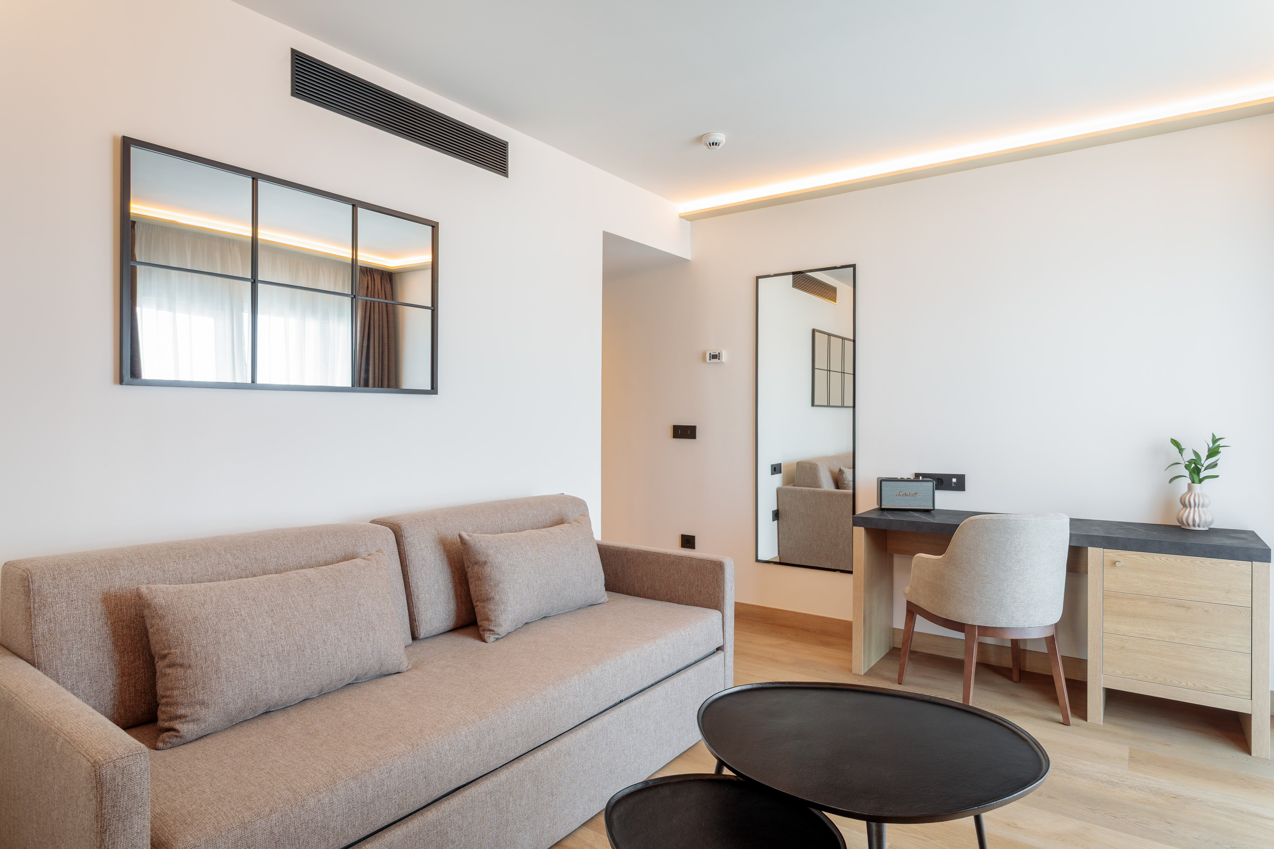 Modern hotel suite with beige sofa, work desk, and decorative mirrors.
