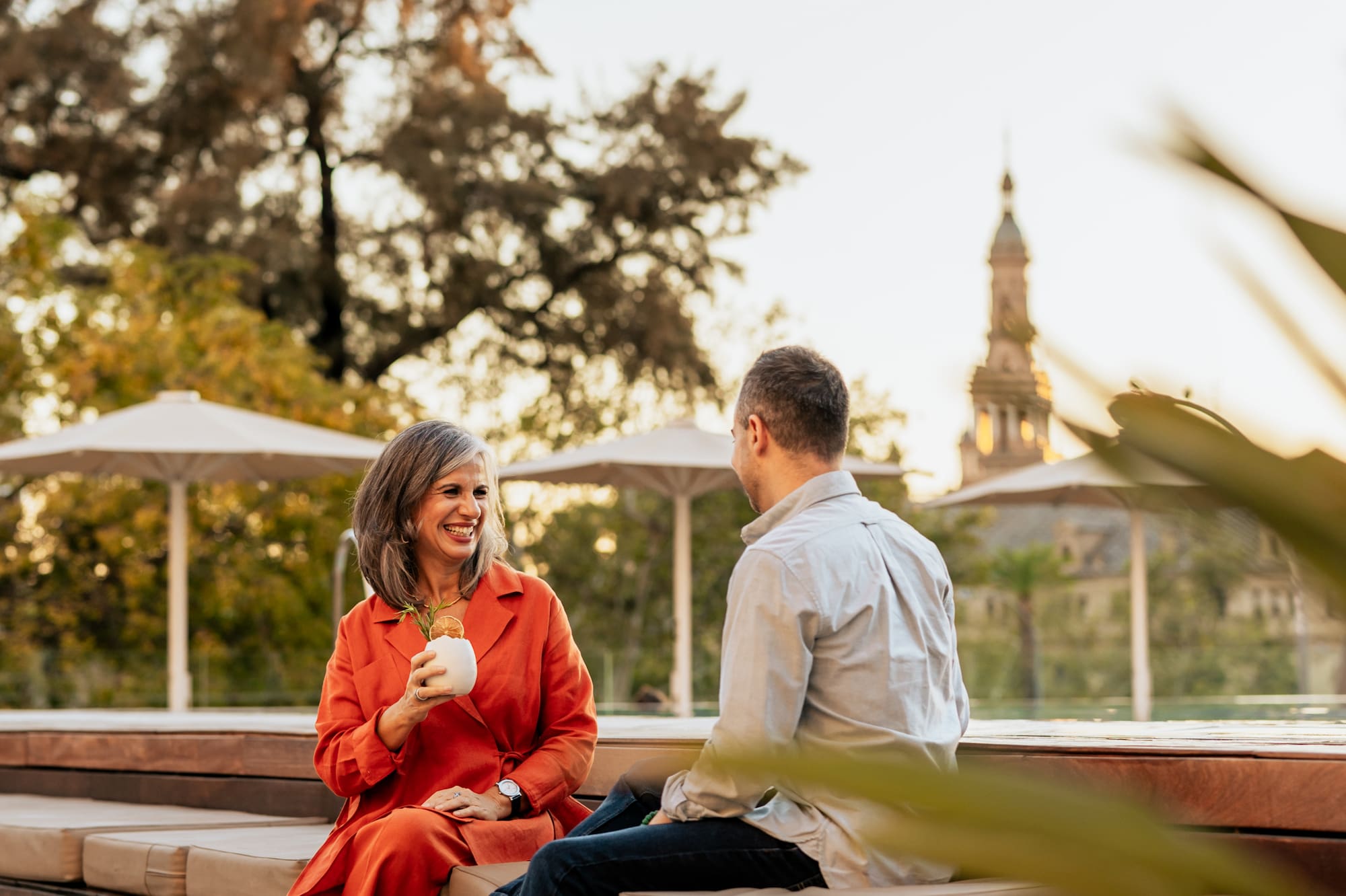 Couple talking on rooftop lounge, woman holding drink, cityscape at golden hour.