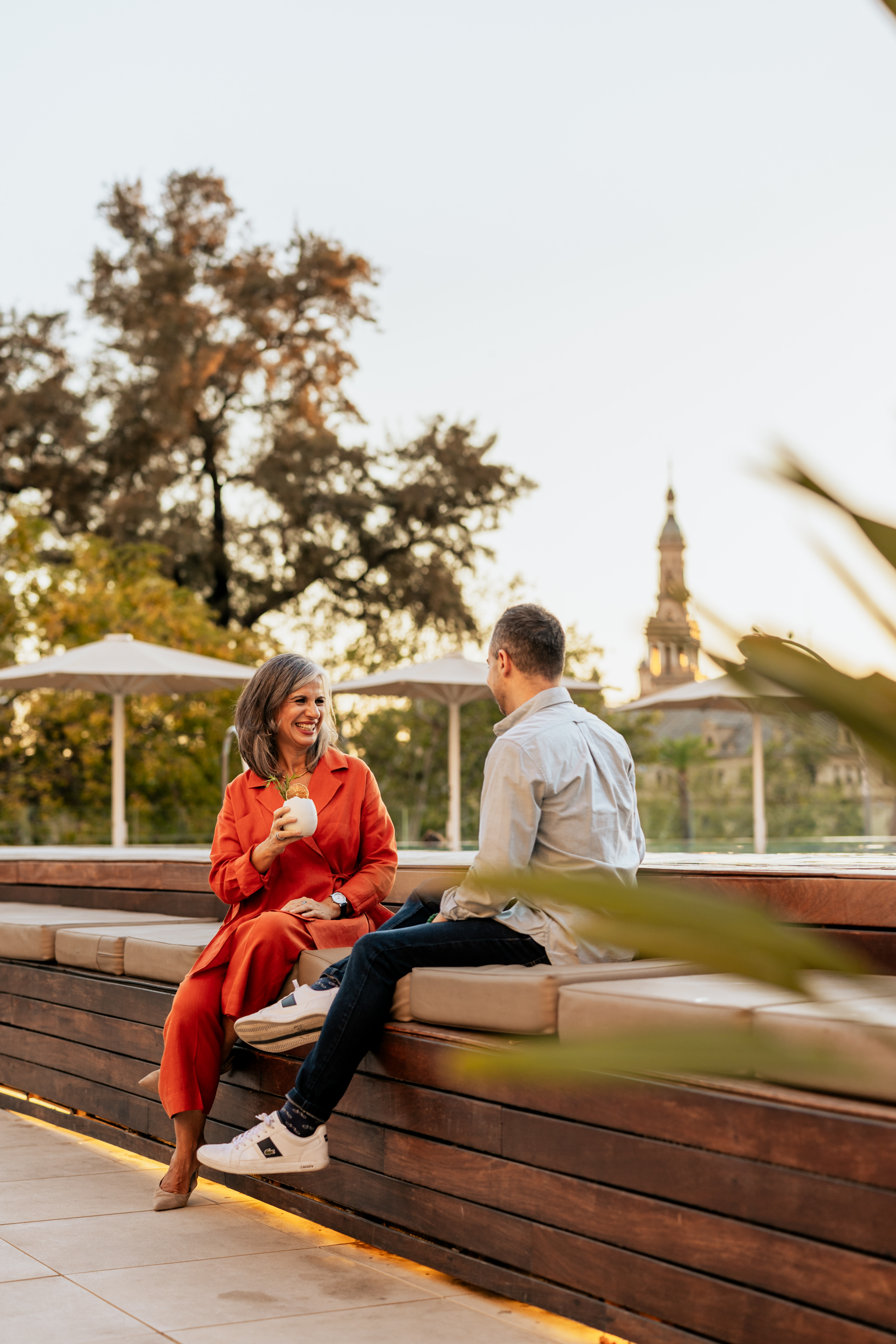 Couple talking on rooftop lounge, woman holding drink, cityscape at golden hour.
