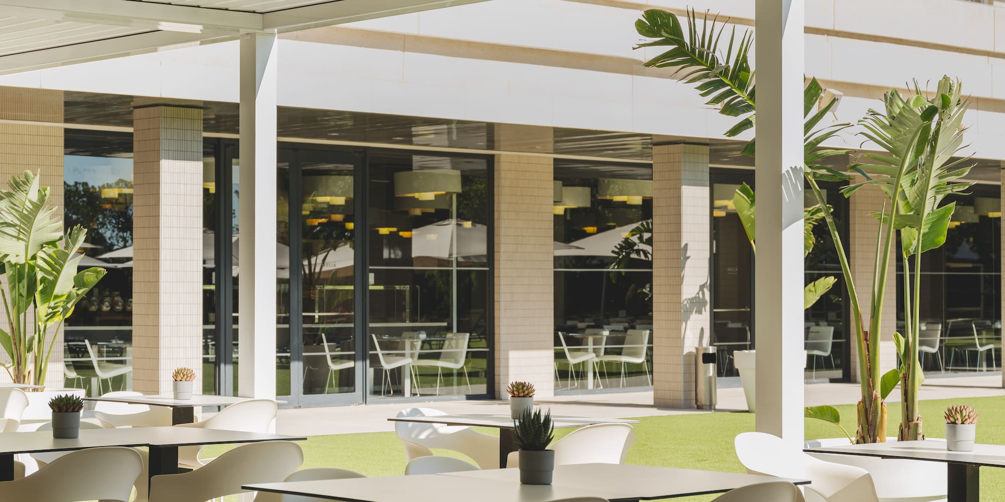 Terraza exterior de restaurante con pérgola, mesas y sillas blancas, césped verde.