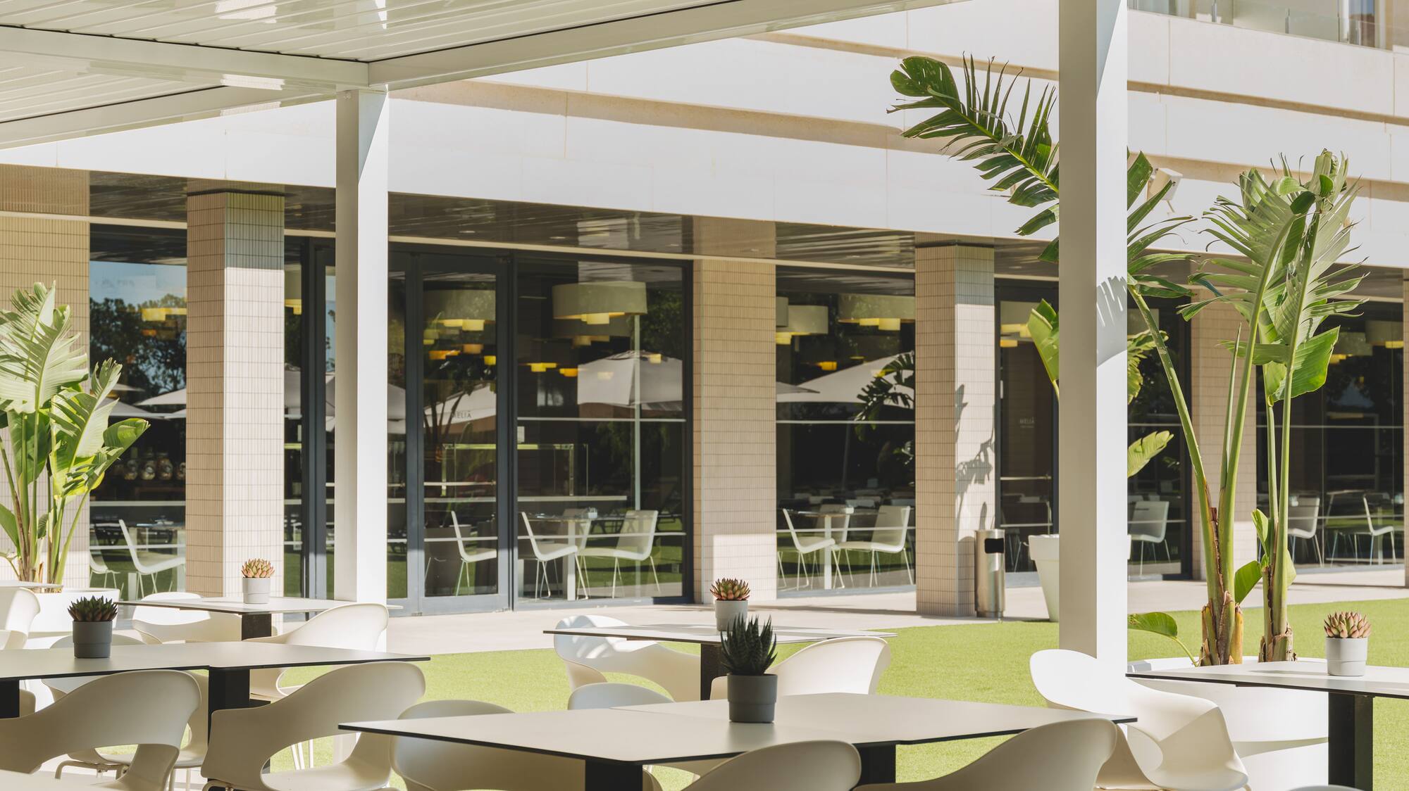Terraza exterior de restaurante con pérgola, mesas y sillas blancas, césped verde.