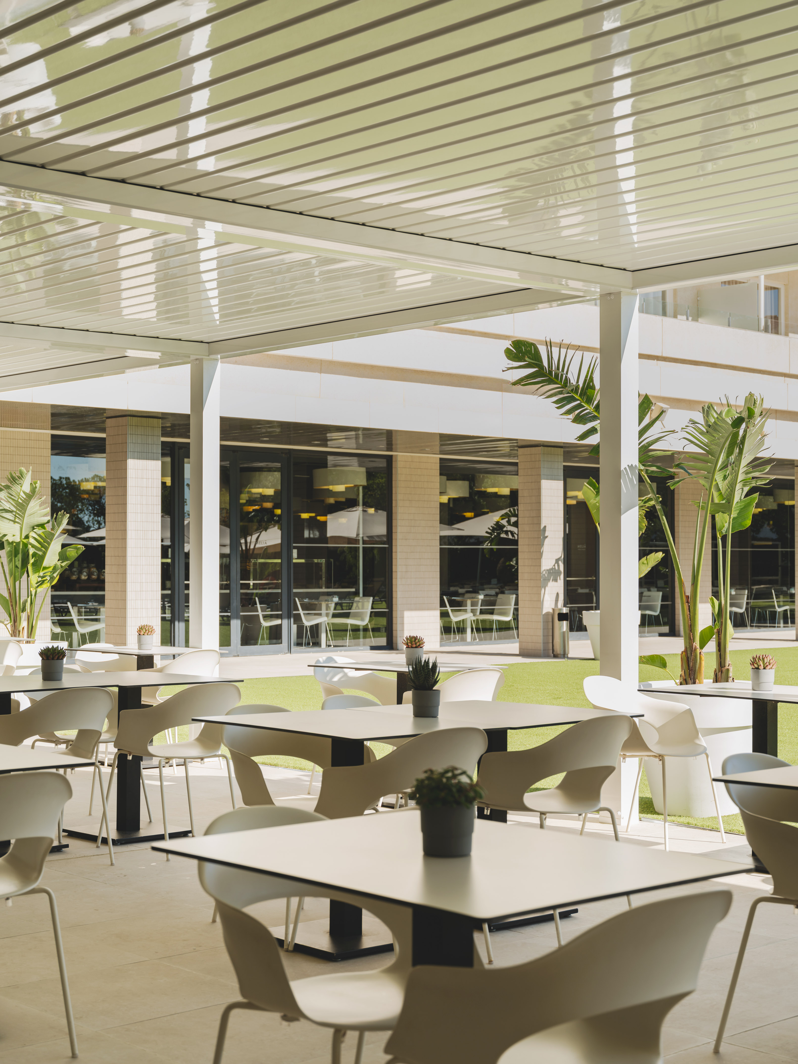 Terraza exterior de restaurante con pérgola, mesas y sillas blancas, césped verde.
