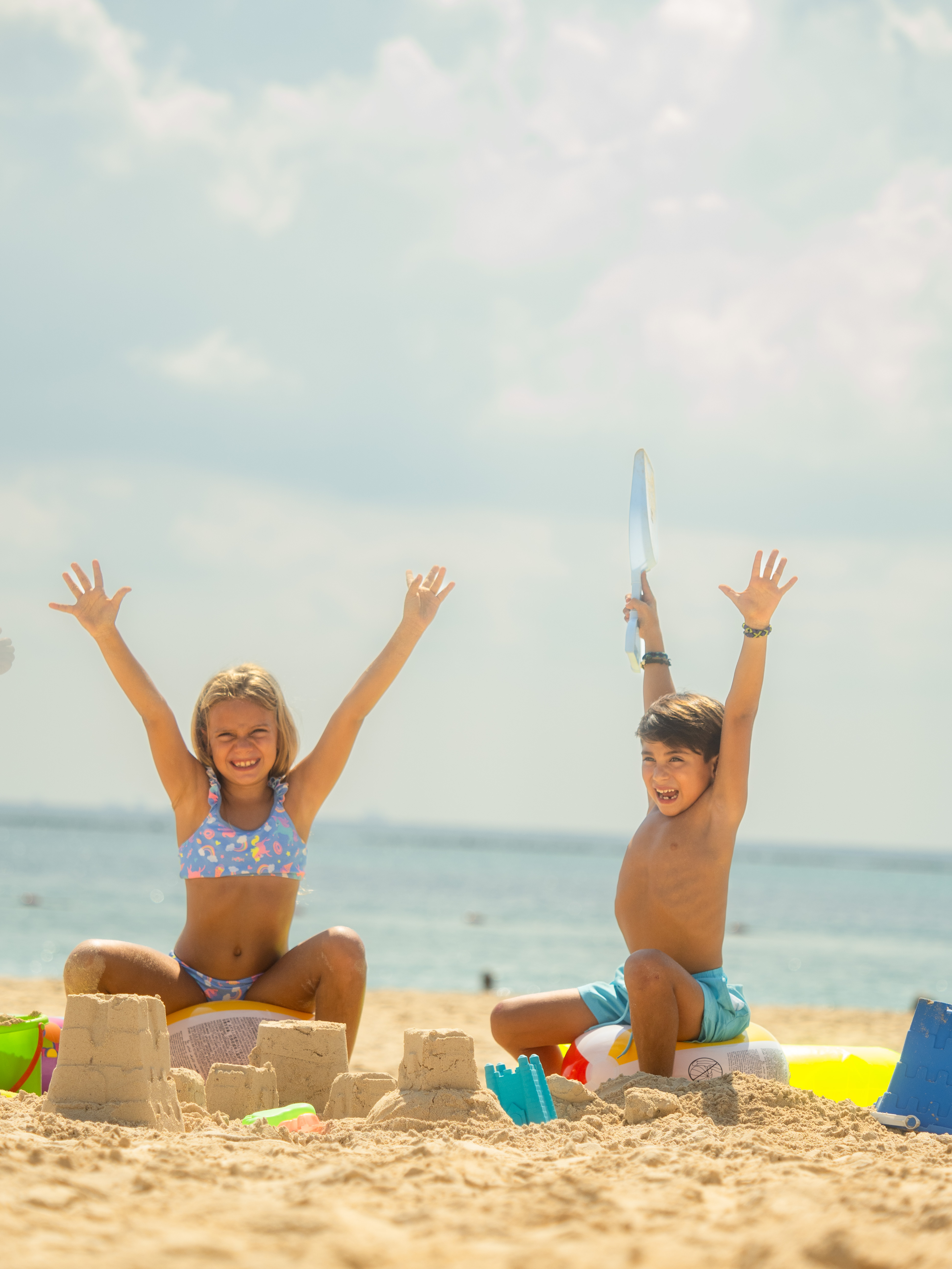 Niños sonrientes jugando en la playa con castillos de arena y mar de fondo.