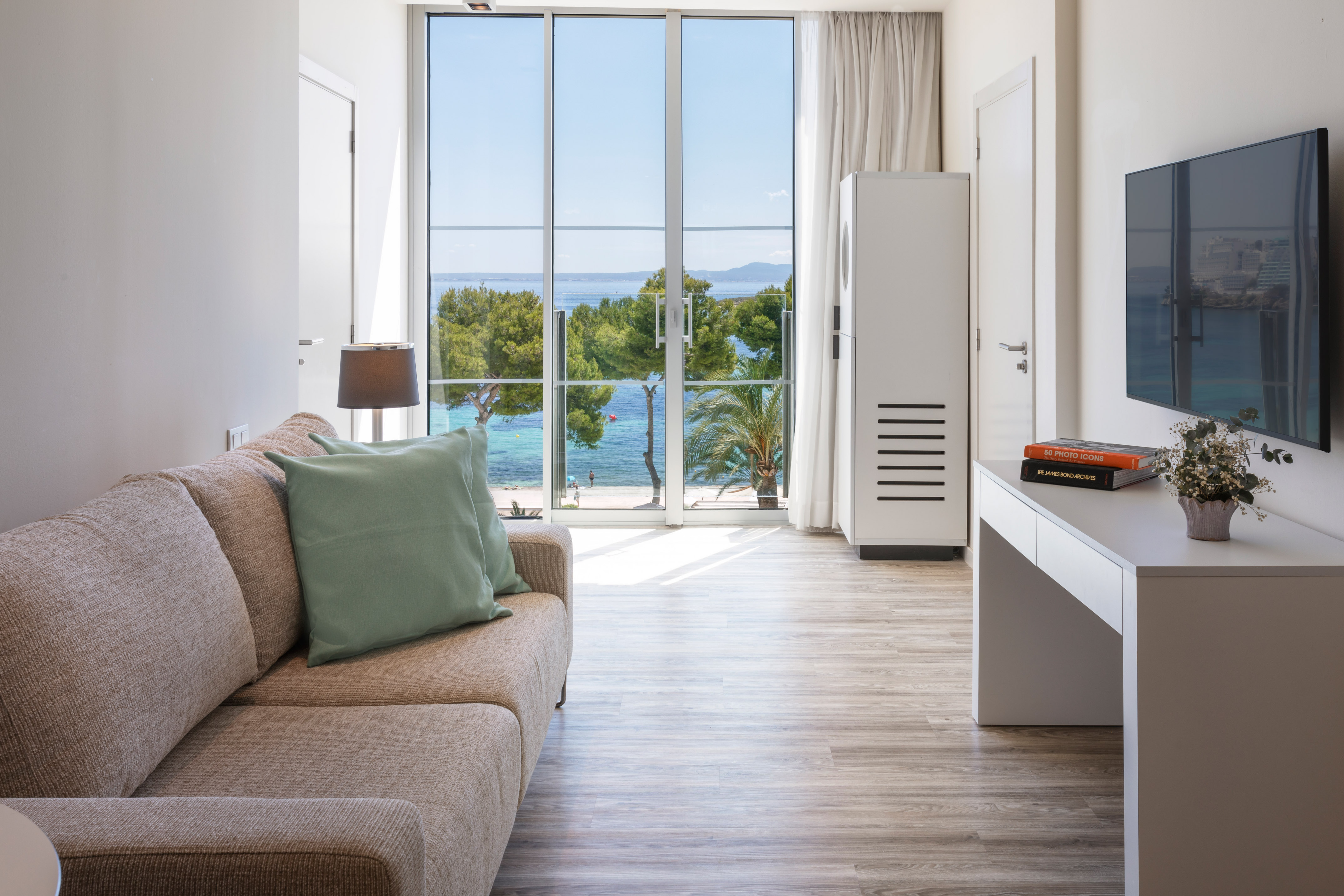 Bright hotel living room with sofa, TV, and panoramic beach-ocean view.