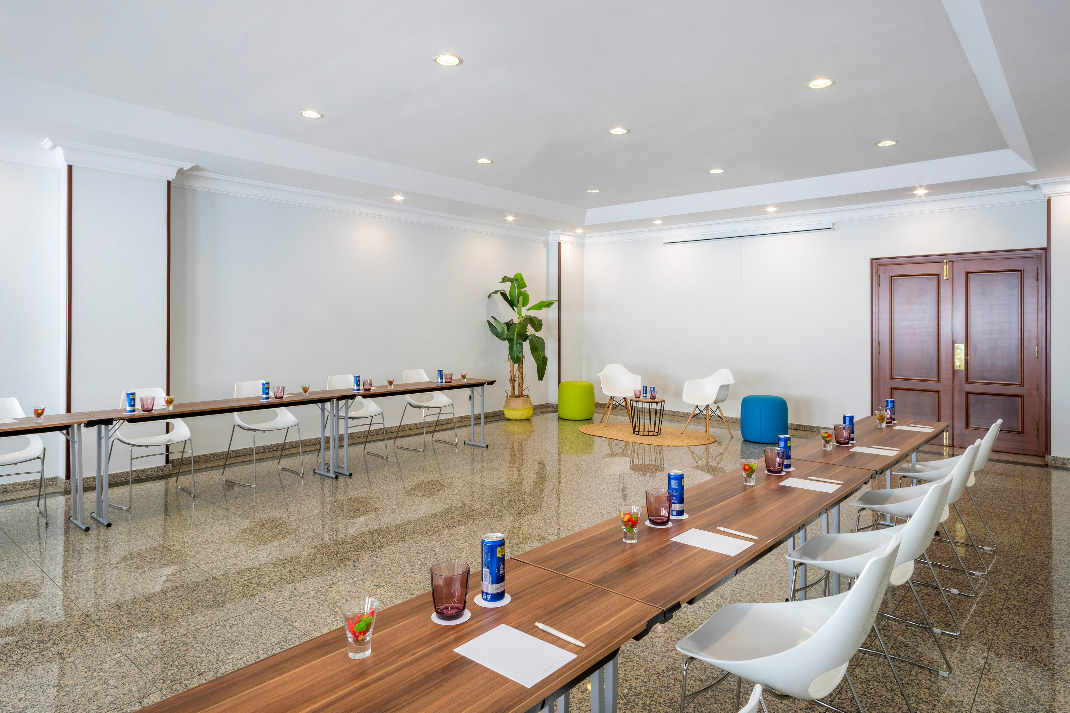 Modern meeting room with U-shaped tables, white chairs, and a small lounge area.