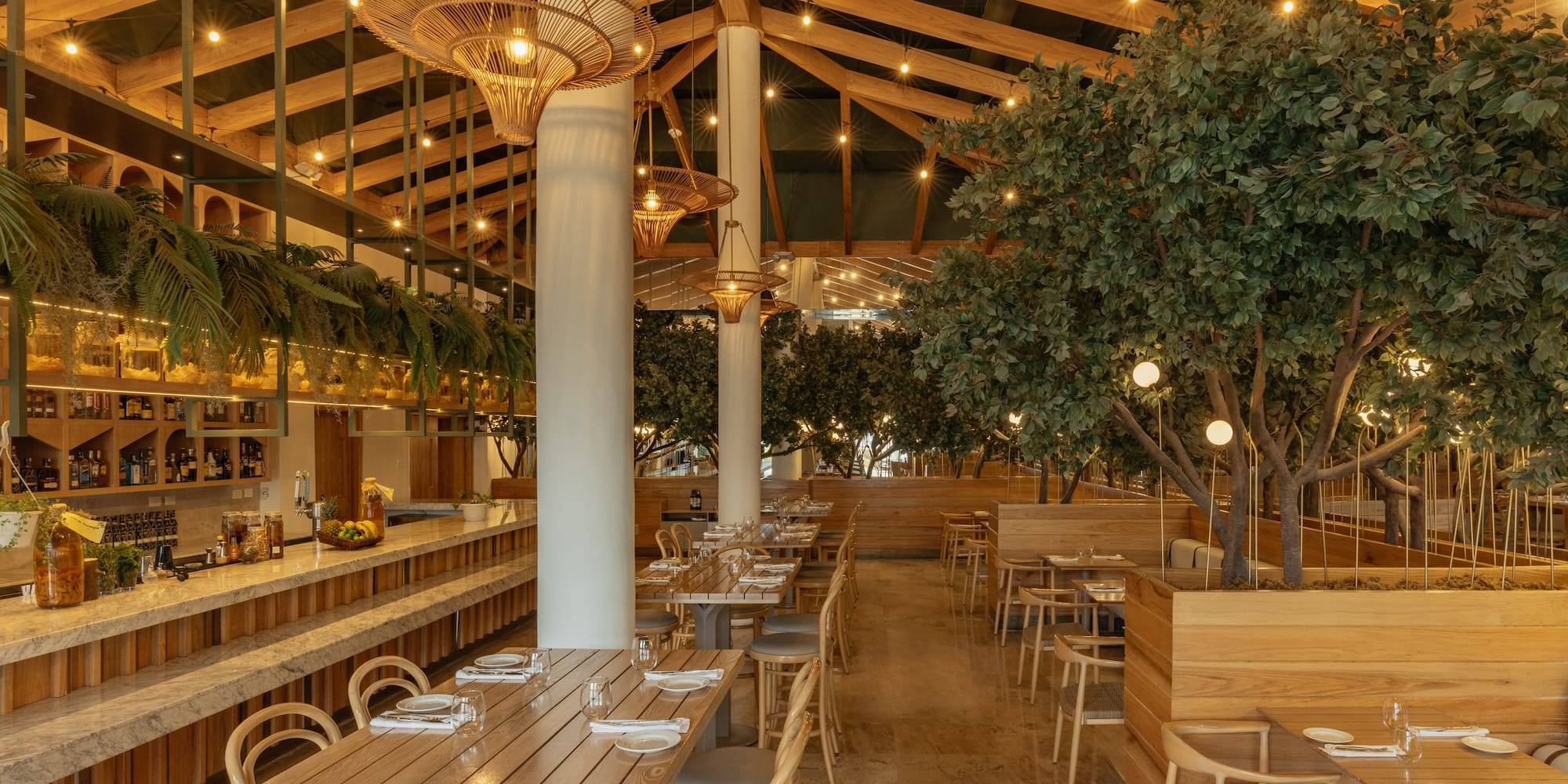 Modern restaurant interior: wooden tables, lush plants, rattan lighting, bar.