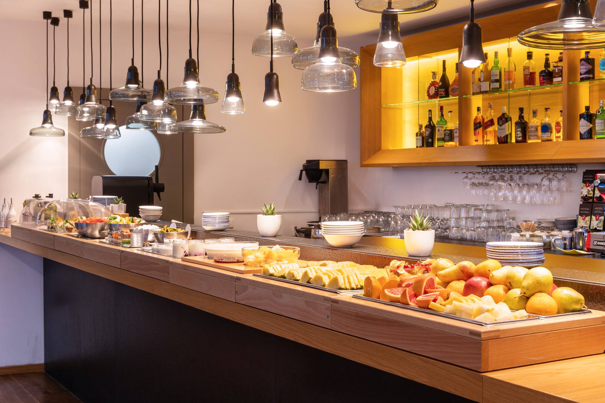 Modern hotel breakfast buffet with fresh fruits, salads, and pendant lights.