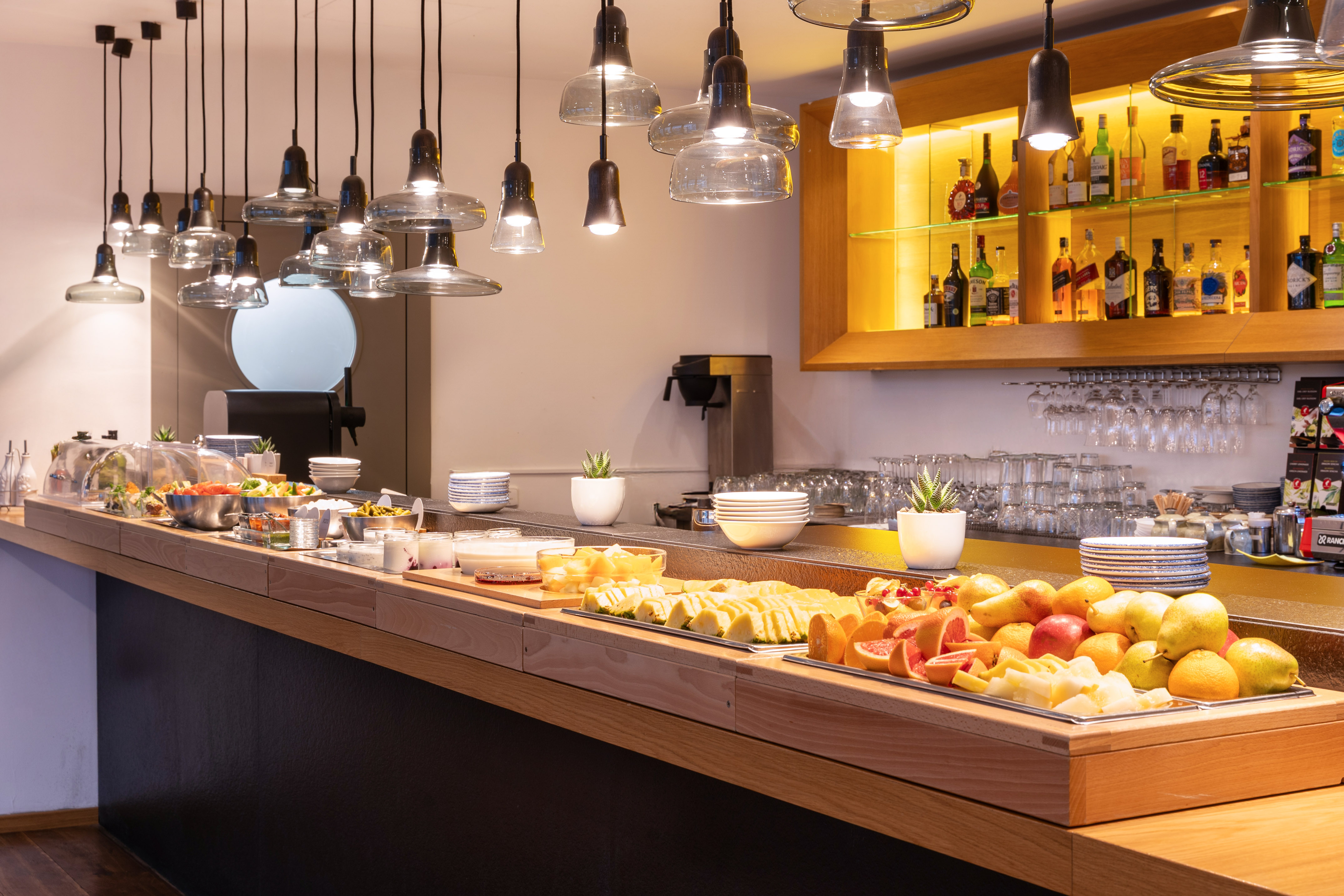 Modern hotel breakfast buffet with fresh fruits, salads, and pendant lights.