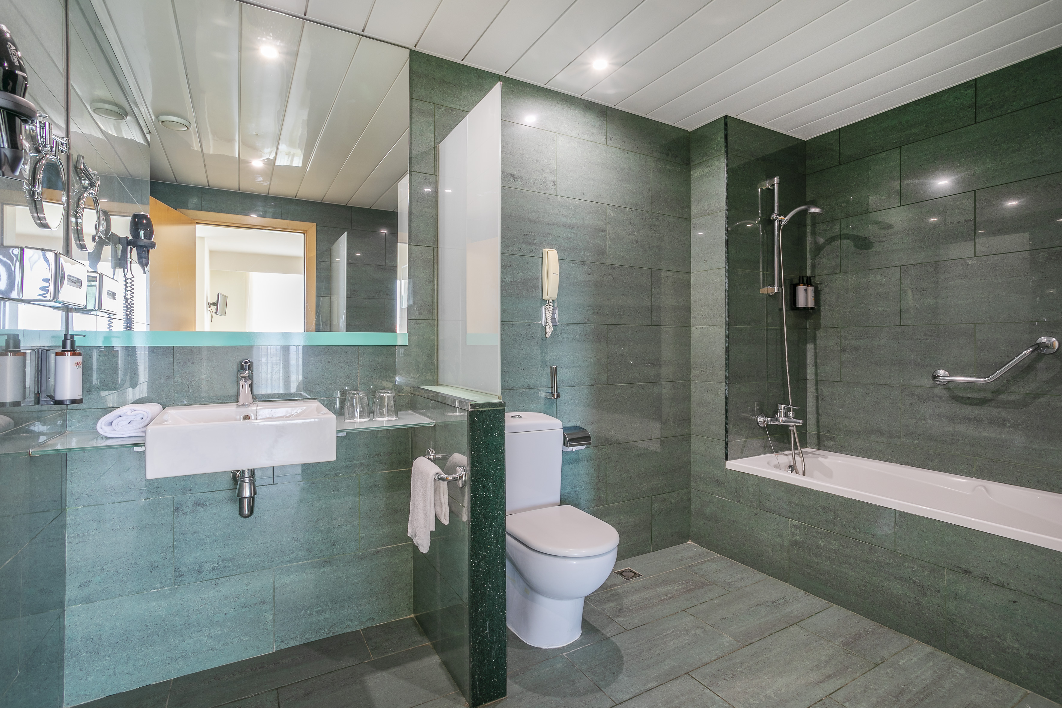 Modern green tiled hotel bathroom with sink, toilet, and bathtub.