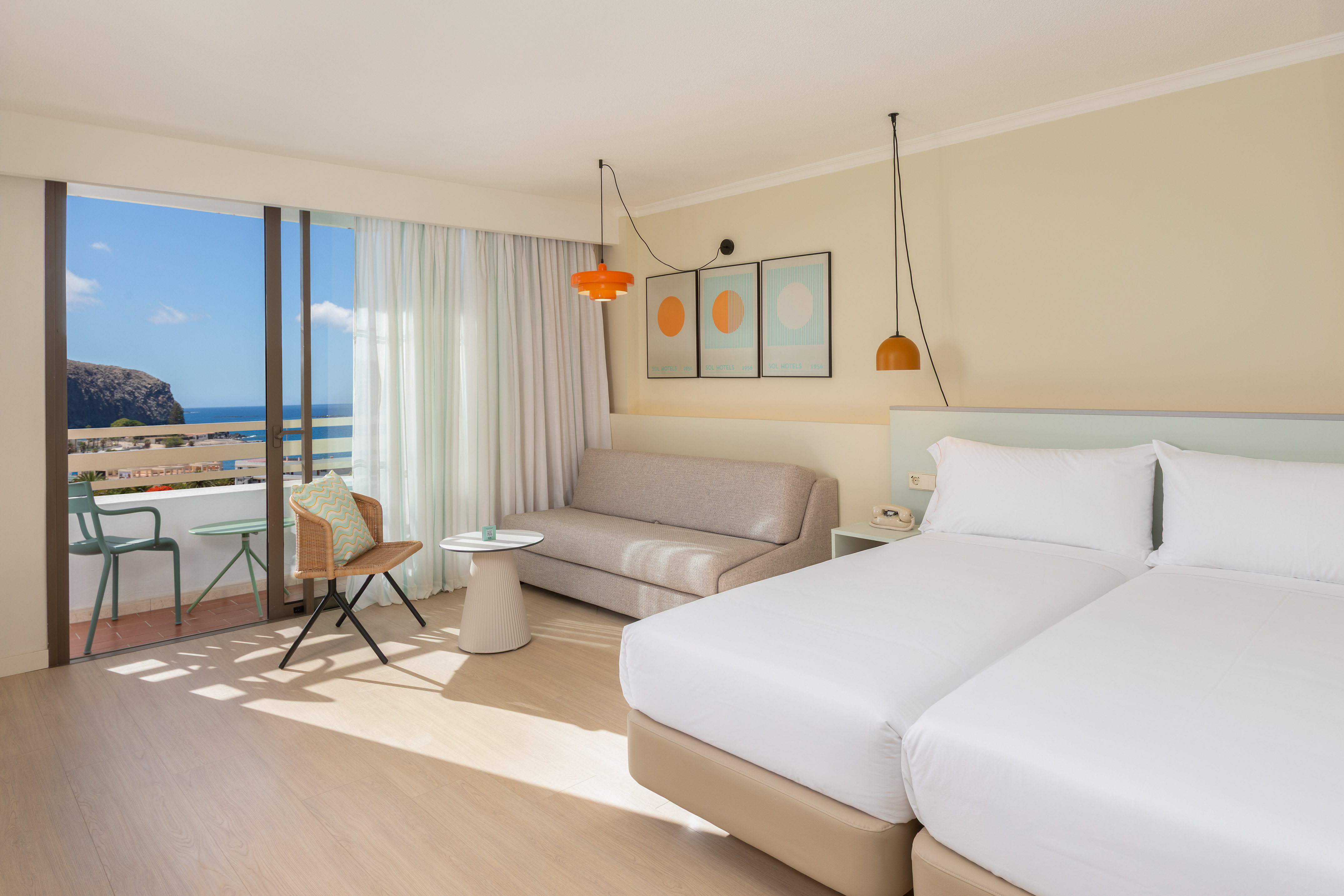 Modern hotel room with twin beds, sofa, and balcony overlooking the sea.