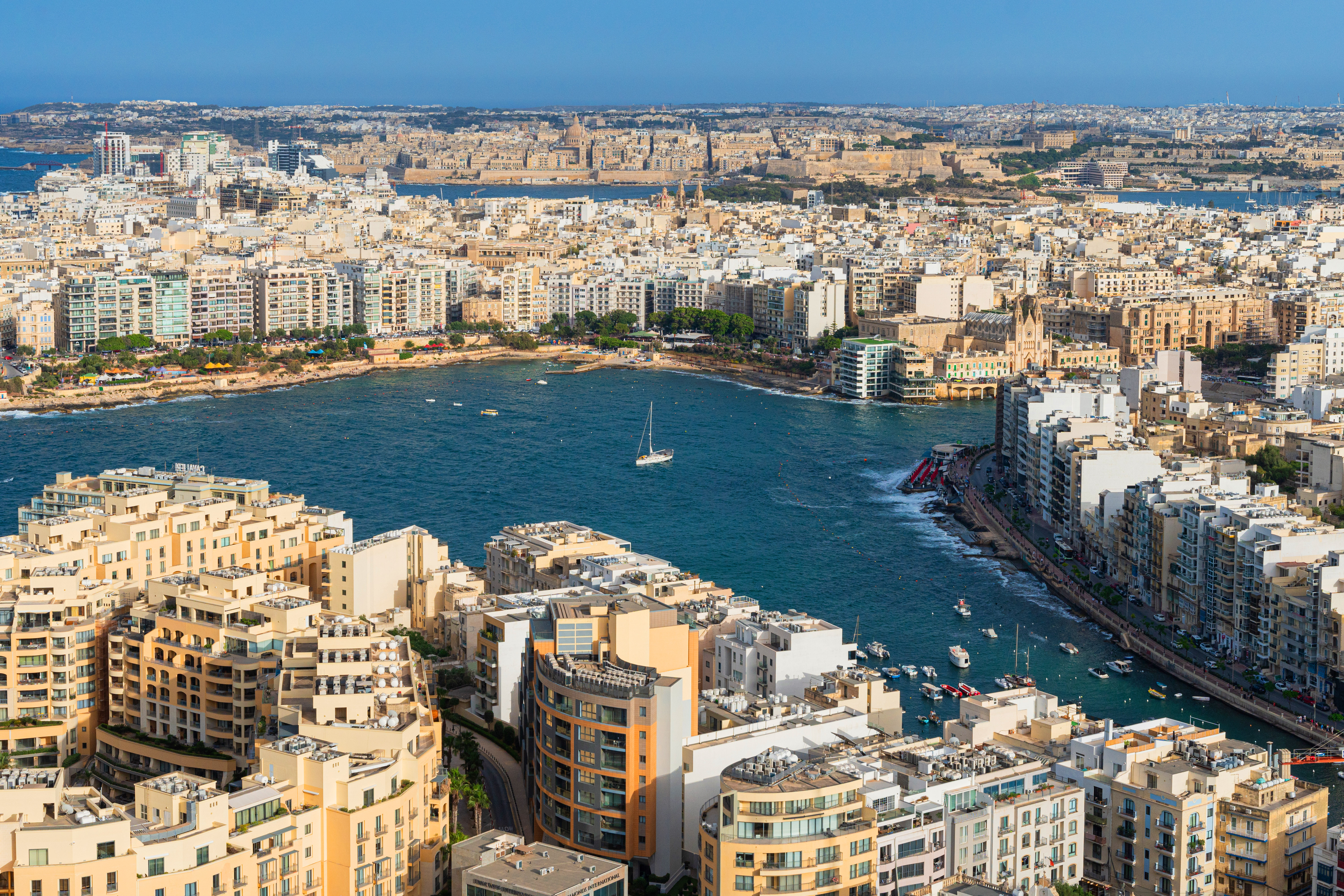 Aerial view of Malta's coastal city, bay with boats, and distant Valletta.