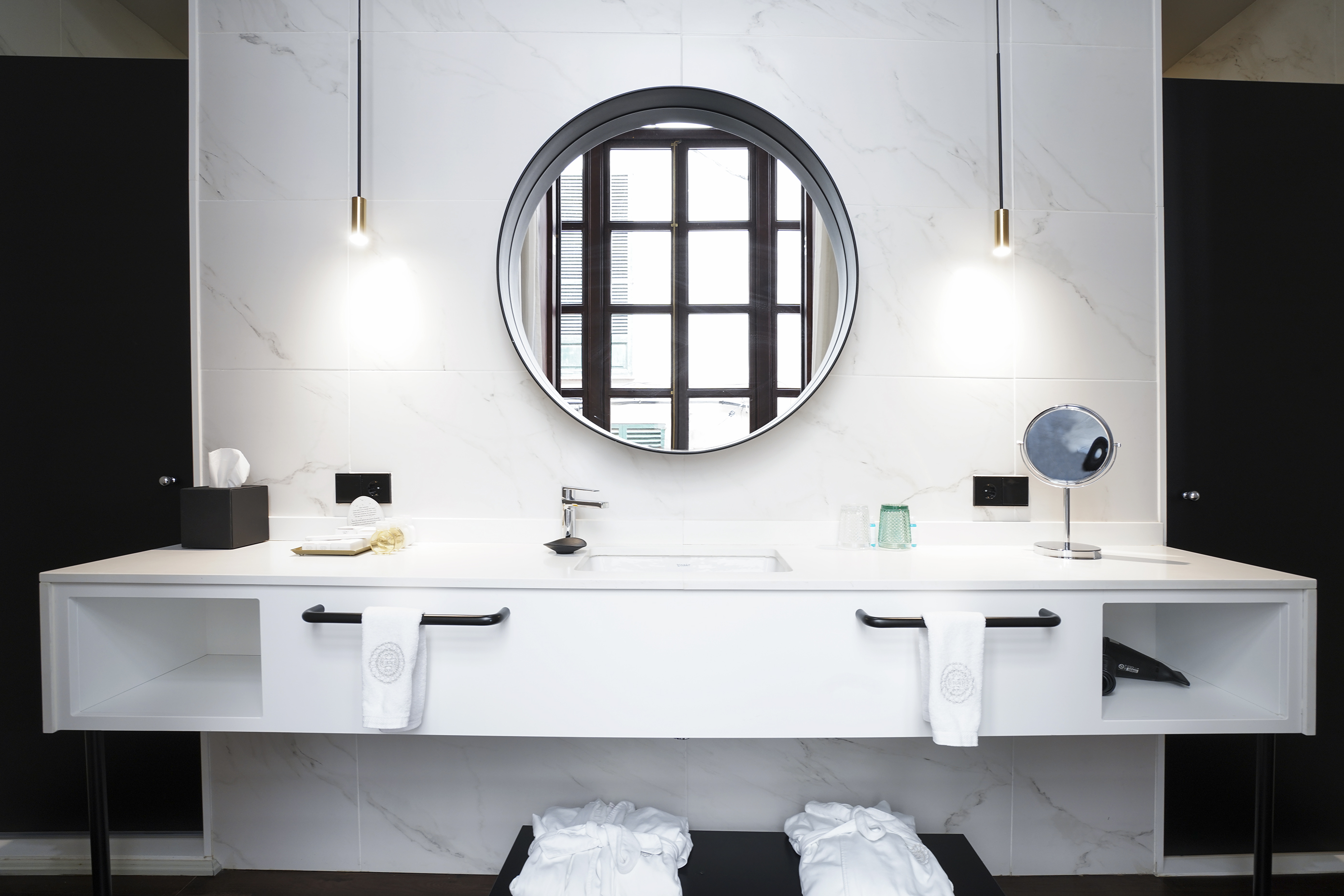 Modern hotel bathroom vanity, round mirror, marble tiles, white sink, pendant lights.