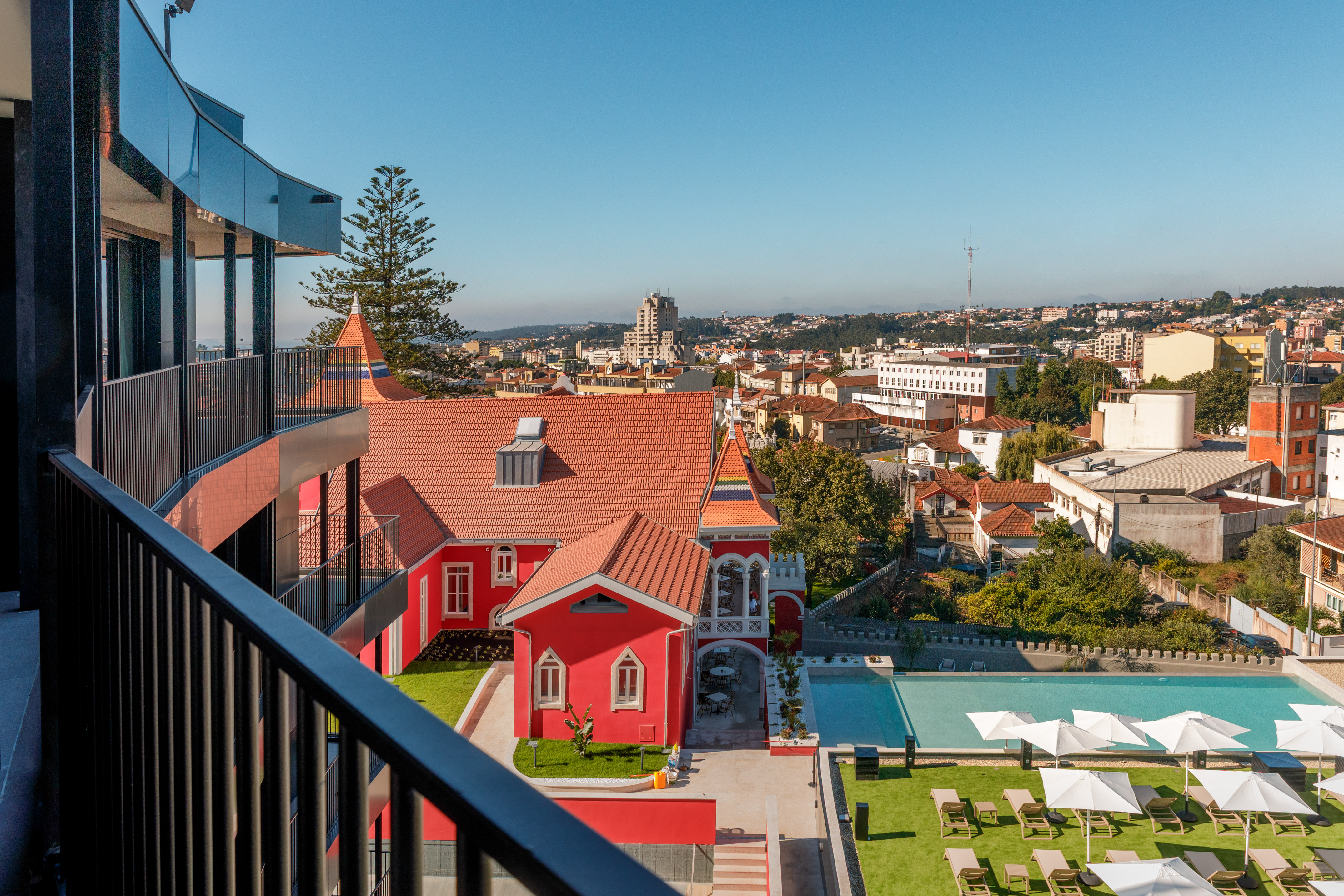 Balcony view overlooking a red hotel, resort swimming pool, and city skyline.