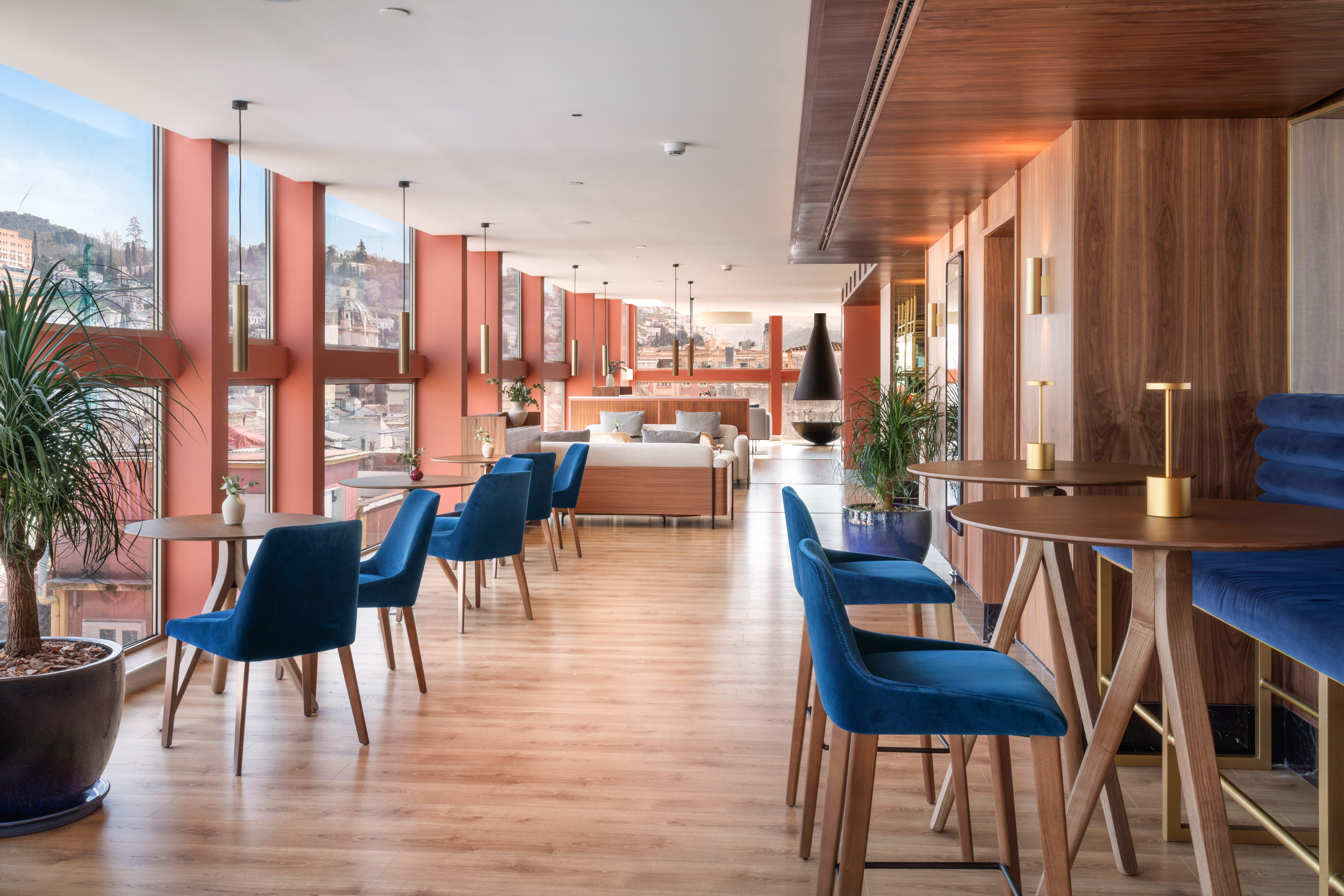 Modern lounge interior with blue velvet chairs, wood tables, city view.