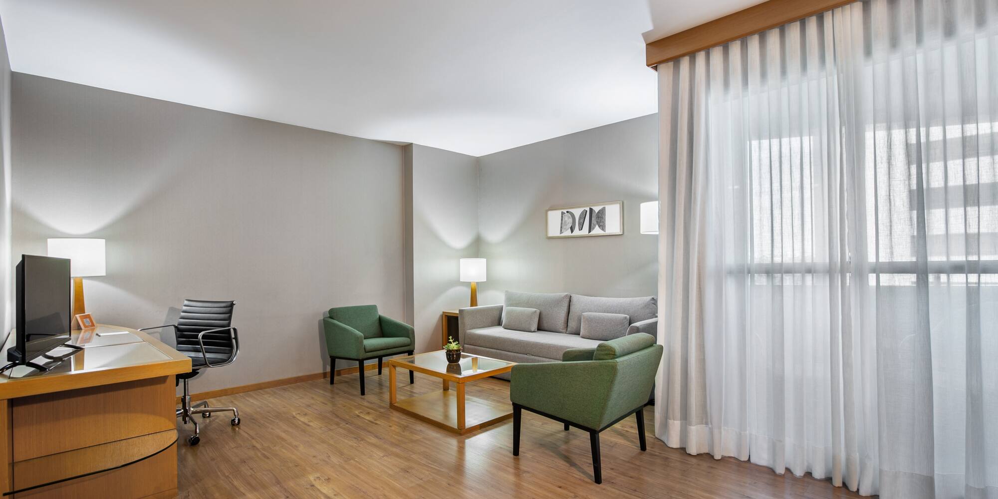 Modern hotel suite with workspace, sofa, green armchairs, and wood floor.