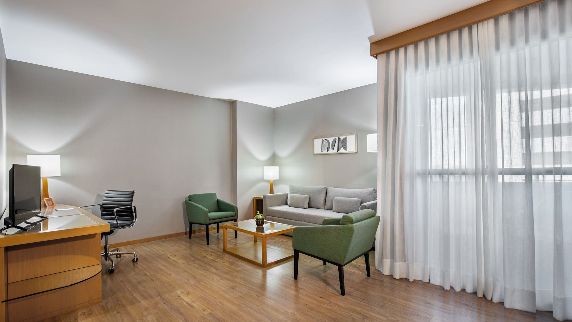 Modern hotel suite with workspace, sofa, green armchairs, and wood floor.