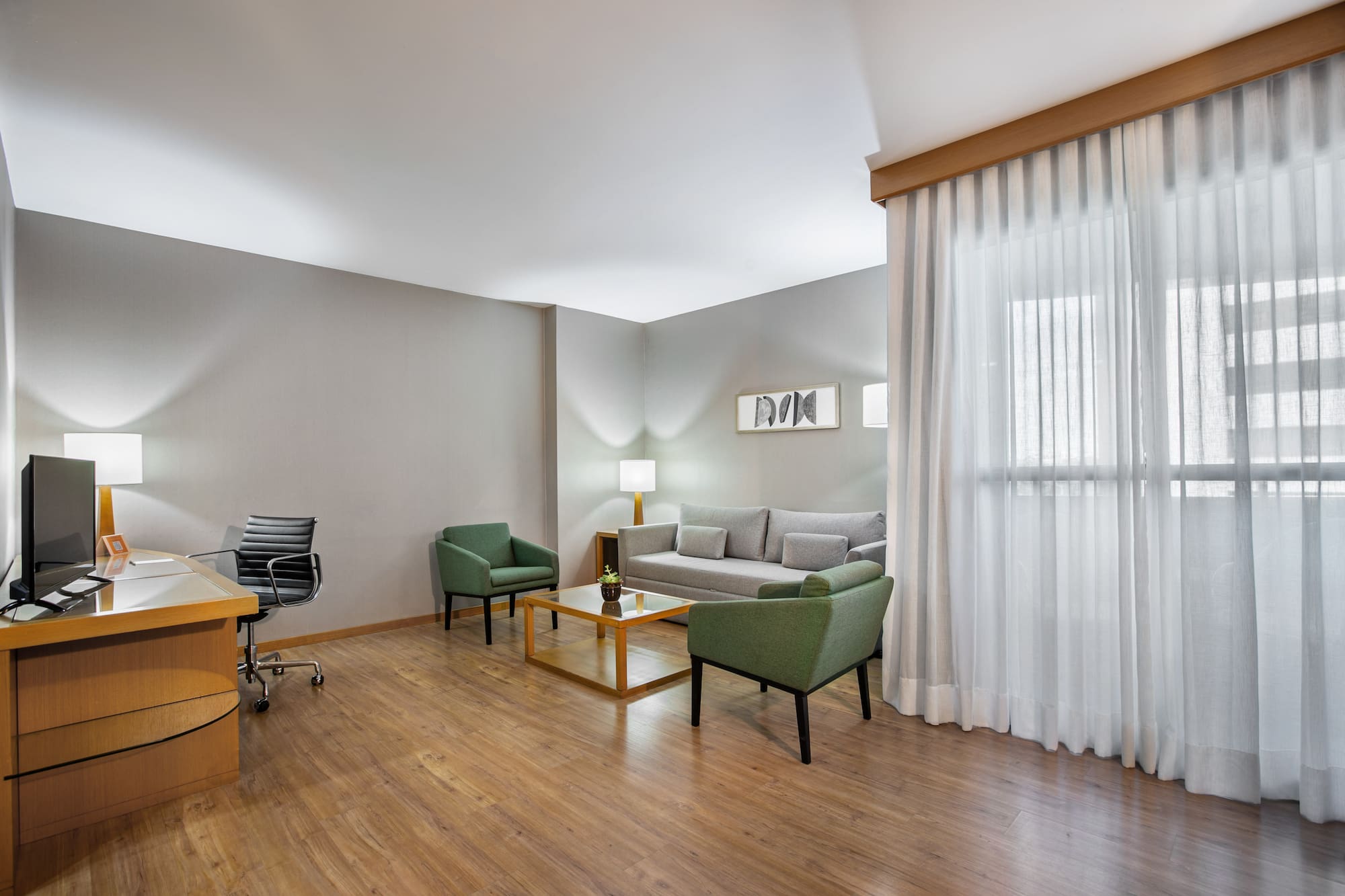 Modern hotel suite with workspace, sofa, green armchairs, and wood floor.