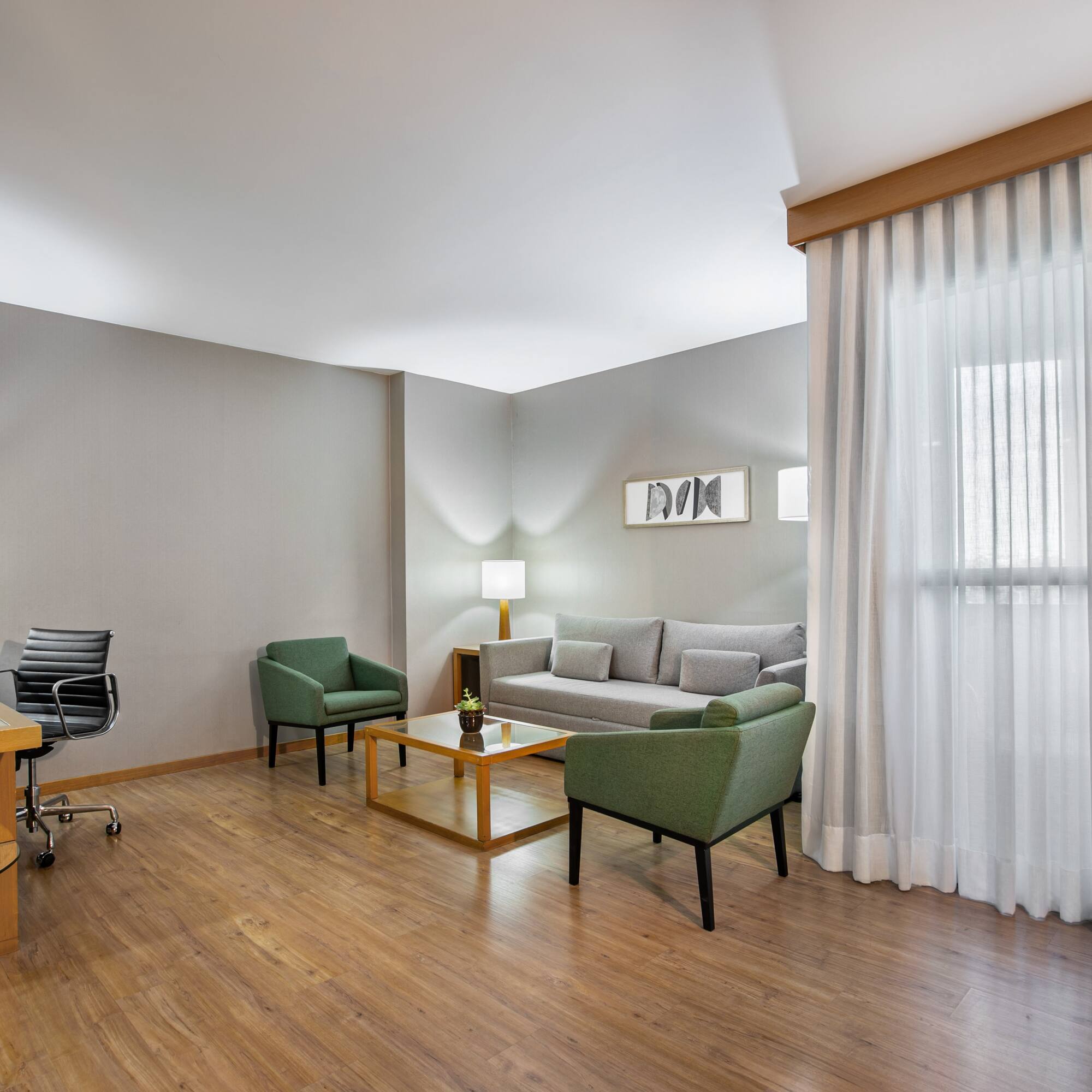 Modern hotel suite with workspace, sofa, green armchairs, and wood floor.