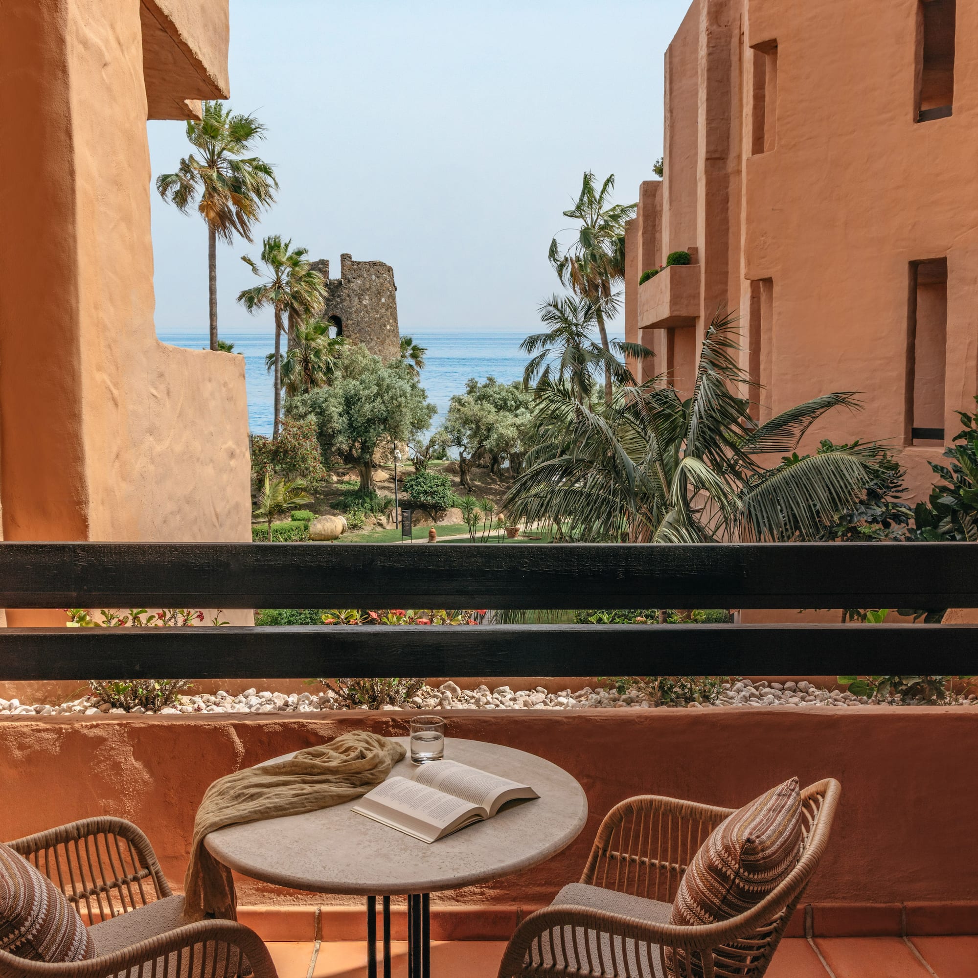 Resort balcony with sea view, palm trees, historic tower, and terracotta architecture.