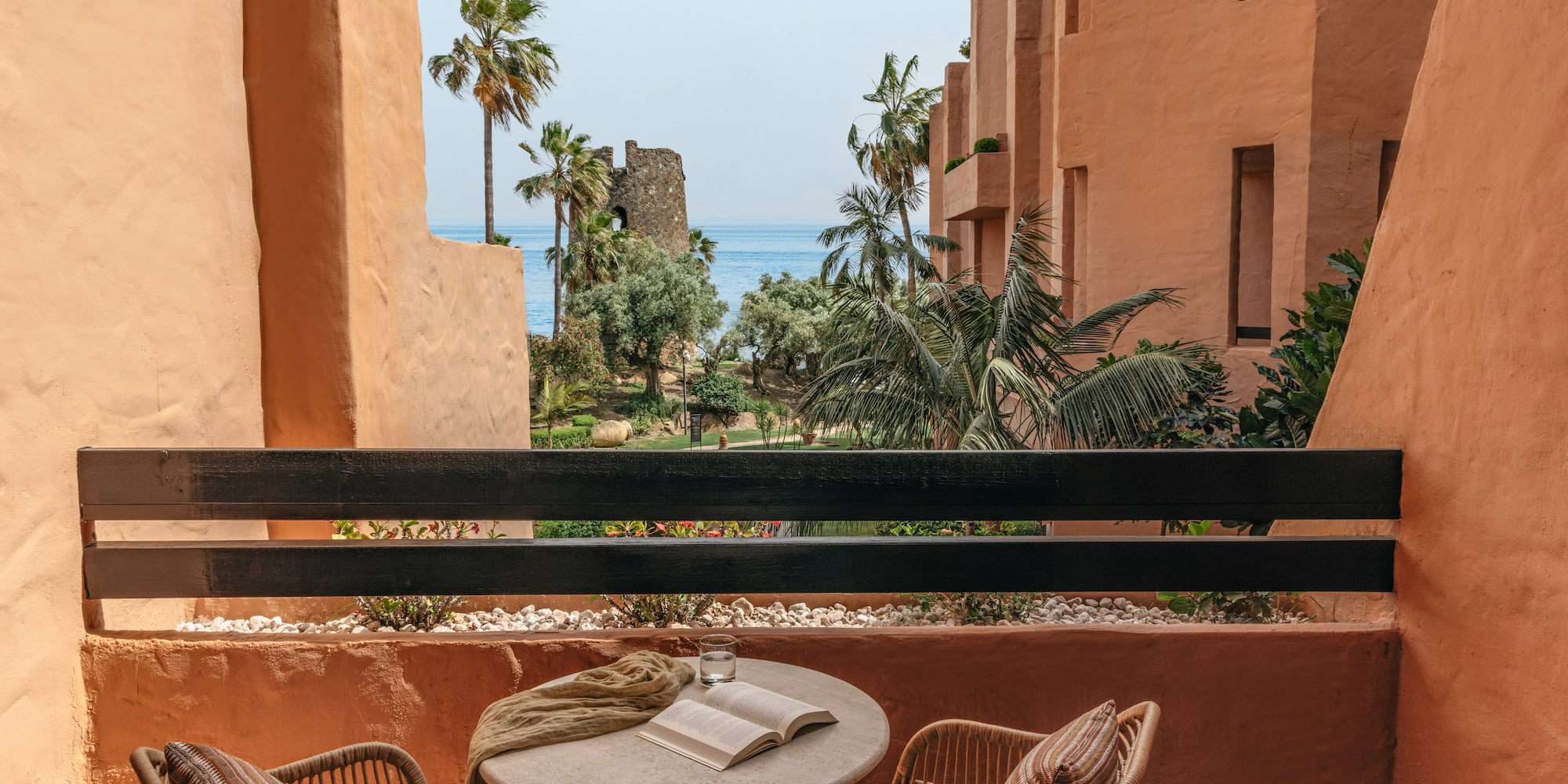 Resort balcony with sea view, palm trees, historic tower, and terracotta architecture.