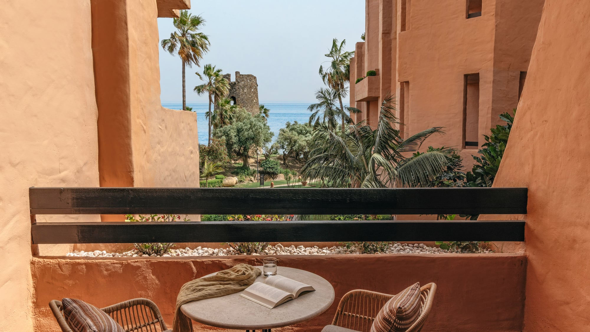 Resort balcony with sea view, palm trees, historic tower, and terracotta architecture.