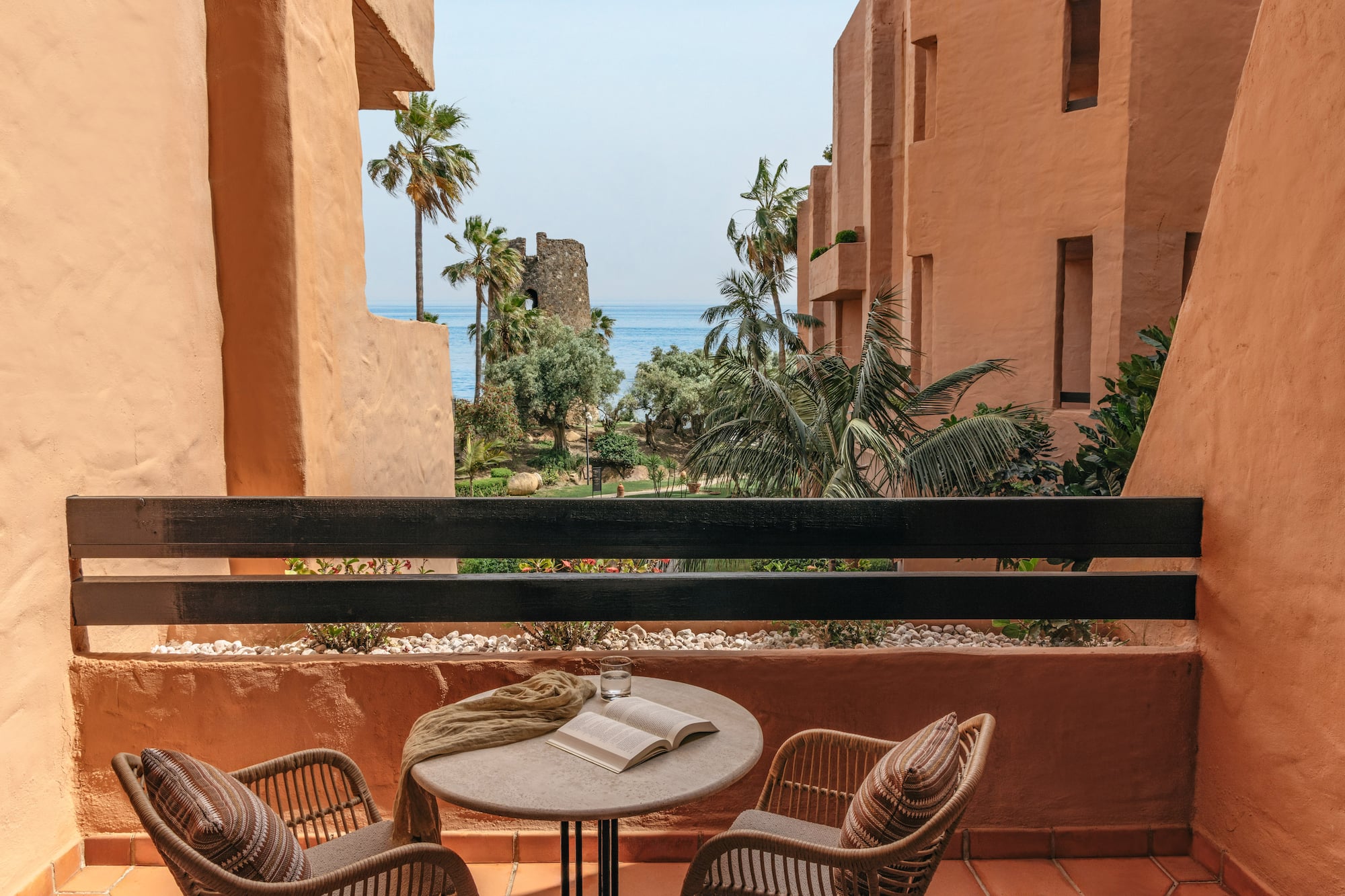 Resort balcony with sea view, palm trees, historic tower, and terracotta architecture.