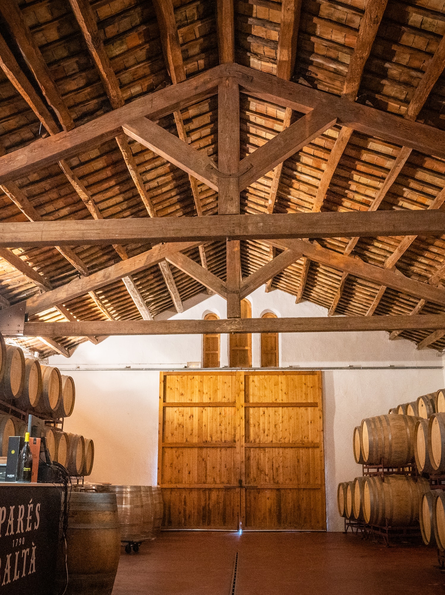 Parés Baltà wine cellar with oak barrels under a rustic wooden beamed ceiling.