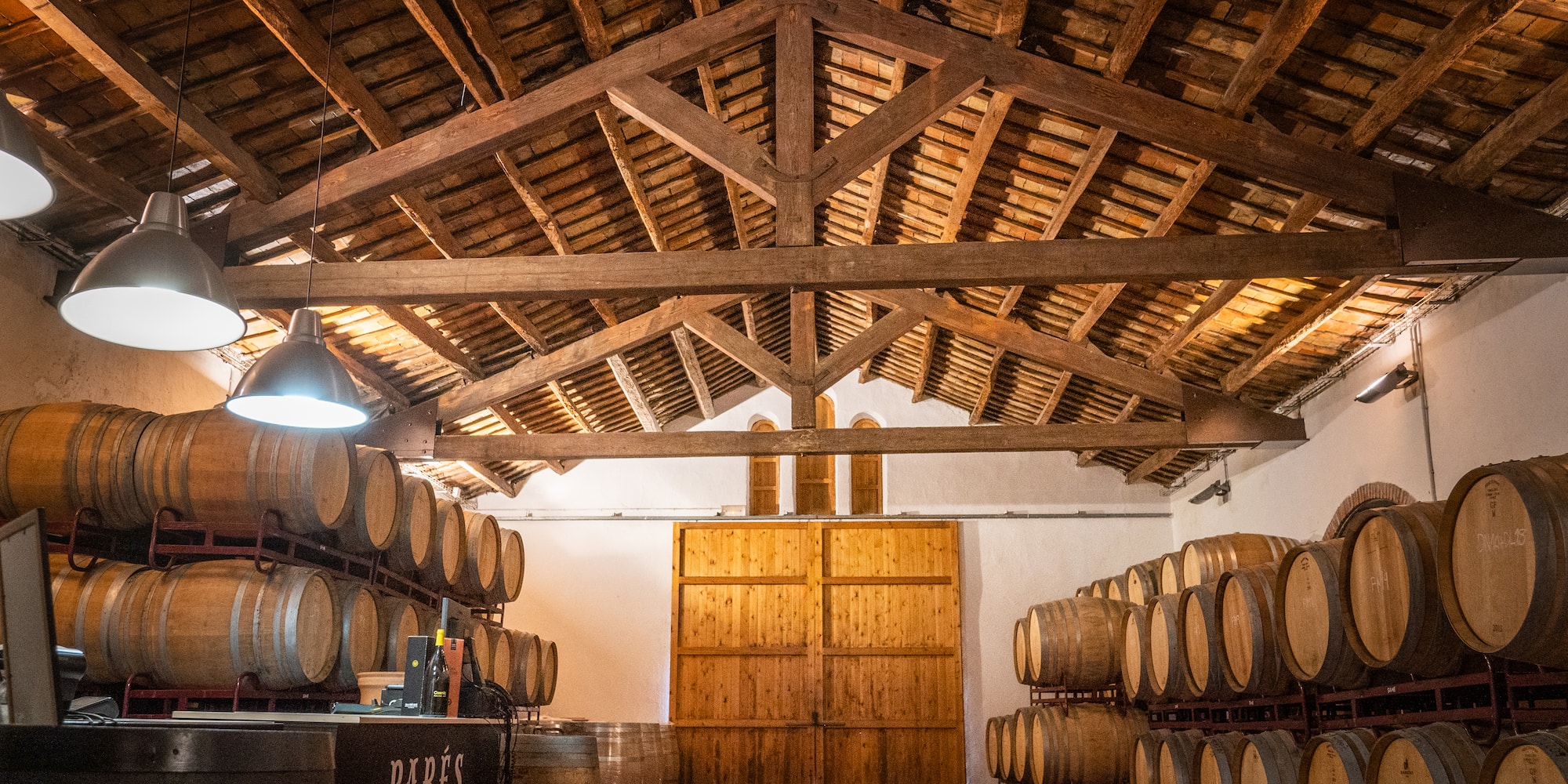 Parés Baltà wine cellar with oak barrels under a rustic wooden beamed ceiling.