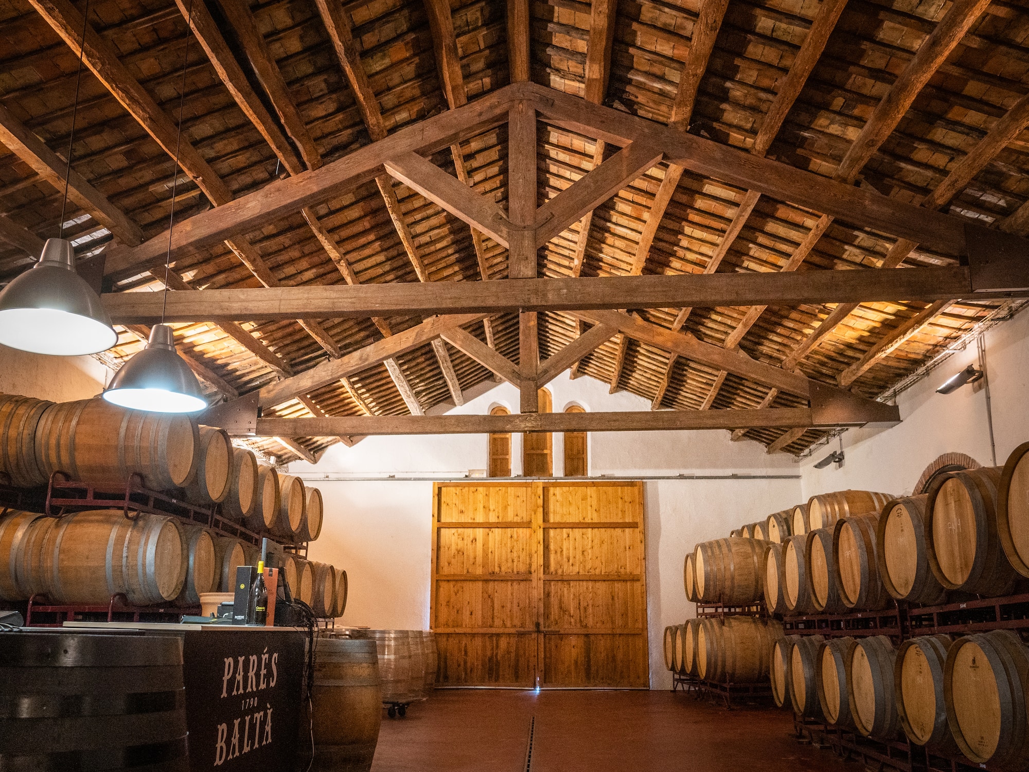Parés Baltà wine cellar with oak barrels under a rustic wooden beamed ceiling.