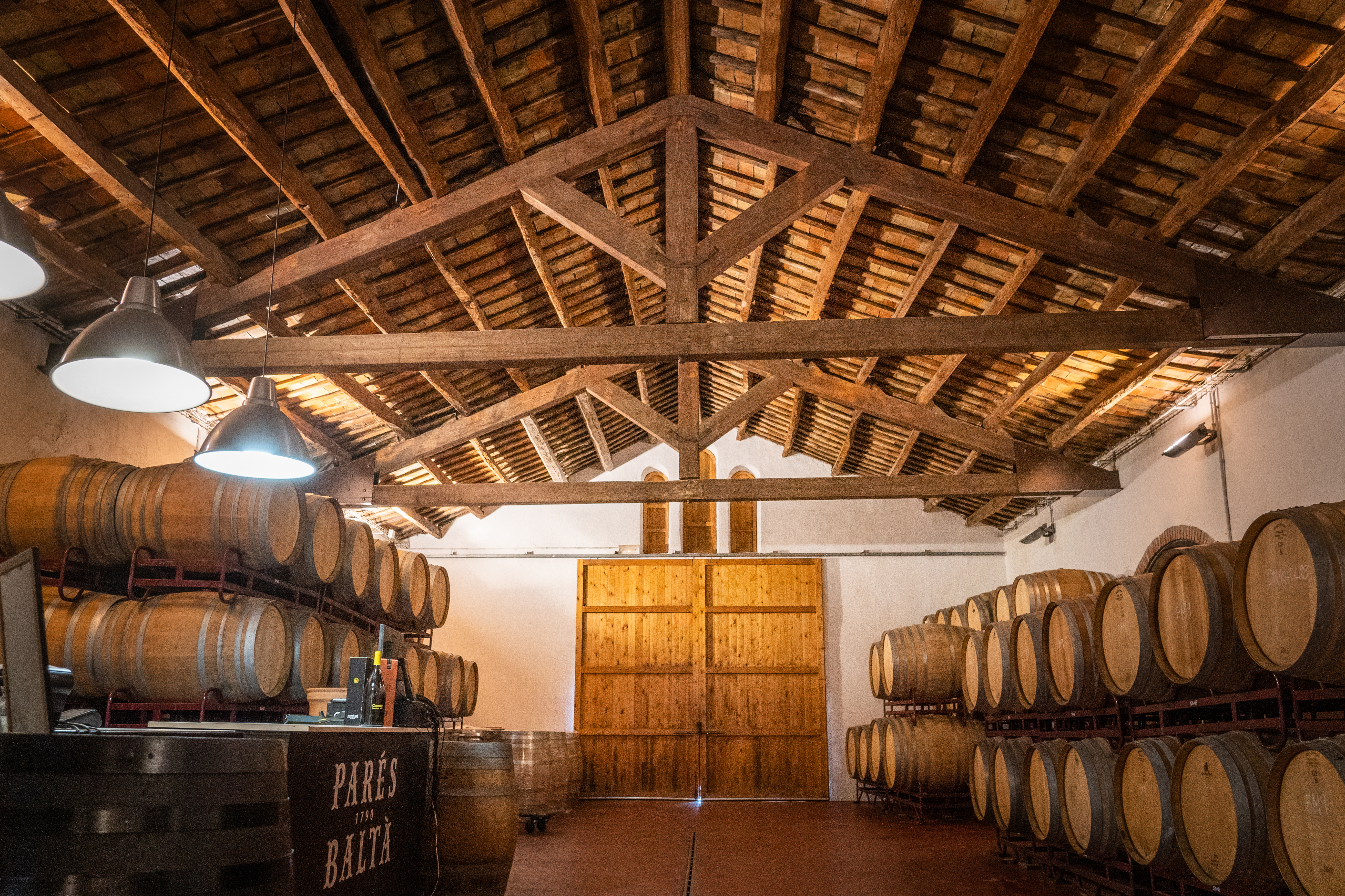 Parés Baltà wine cellar with oak barrels under a rustic wooden beamed ceiling.