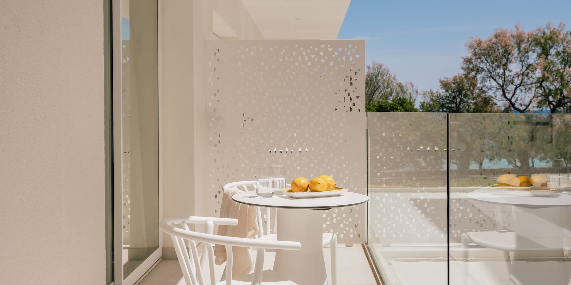 Modern white balcony with outdoor furniture, lemons, glass railing, and green trees.