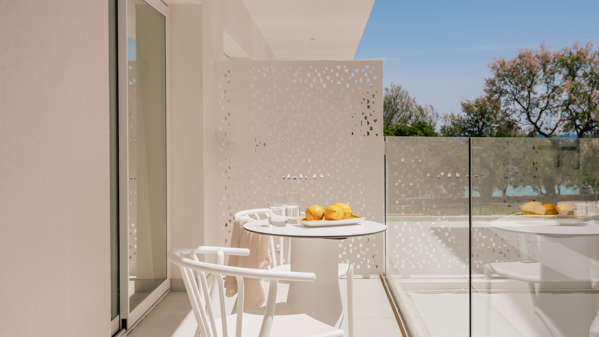 Modern white balcony with outdoor furniture, lemons, glass railing, and green trees.