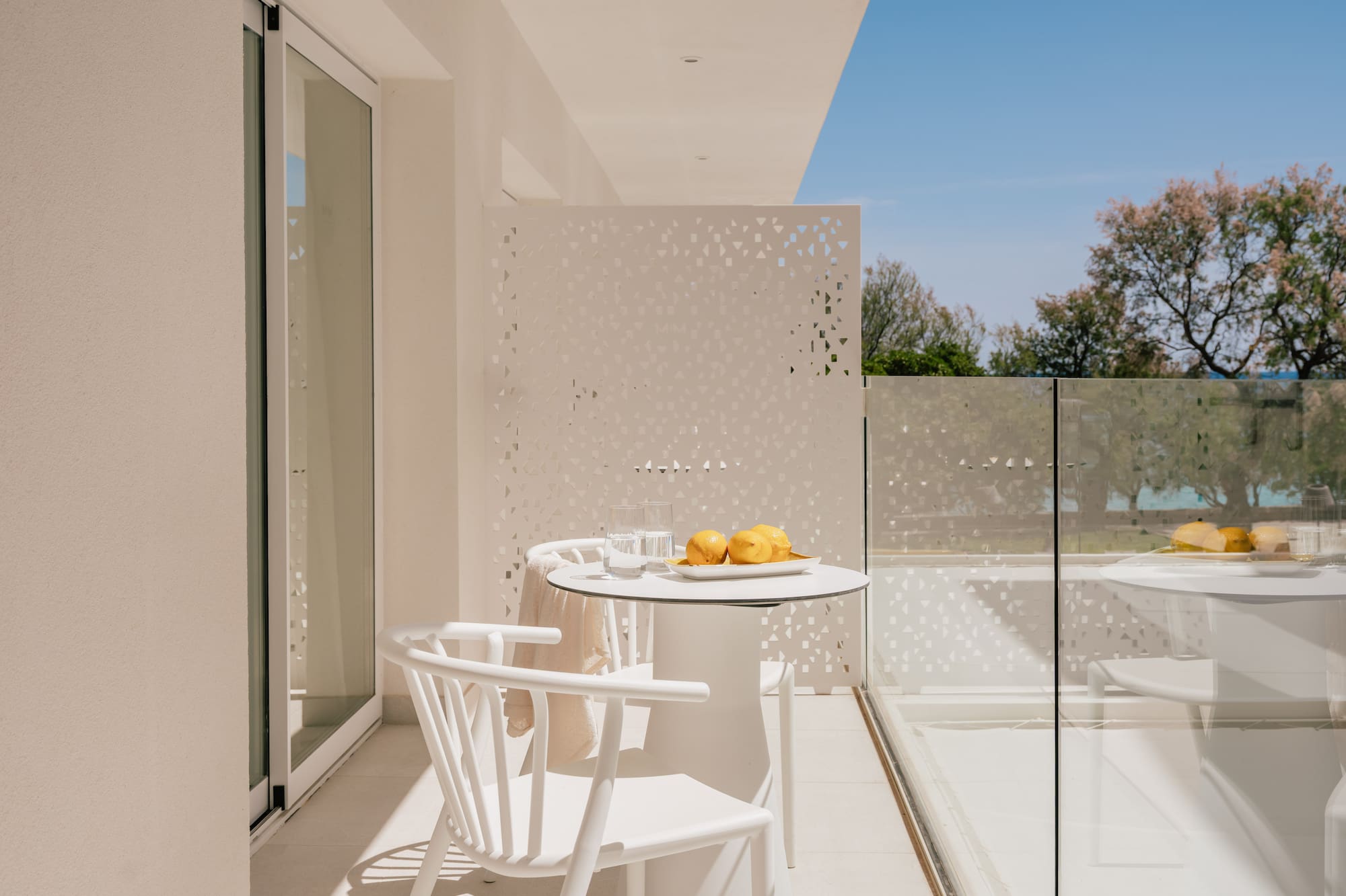 Modern white balcony with outdoor furniture, lemons, glass railing, and green trees.