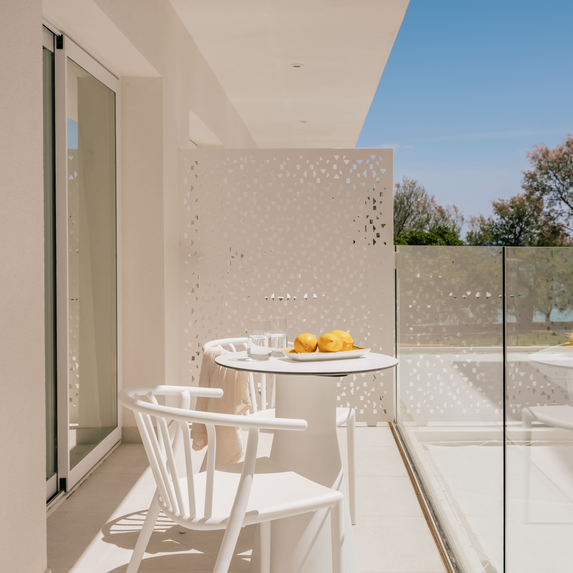 Modern white balcony with outdoor furniture, lemons, glass railing, and green trees.