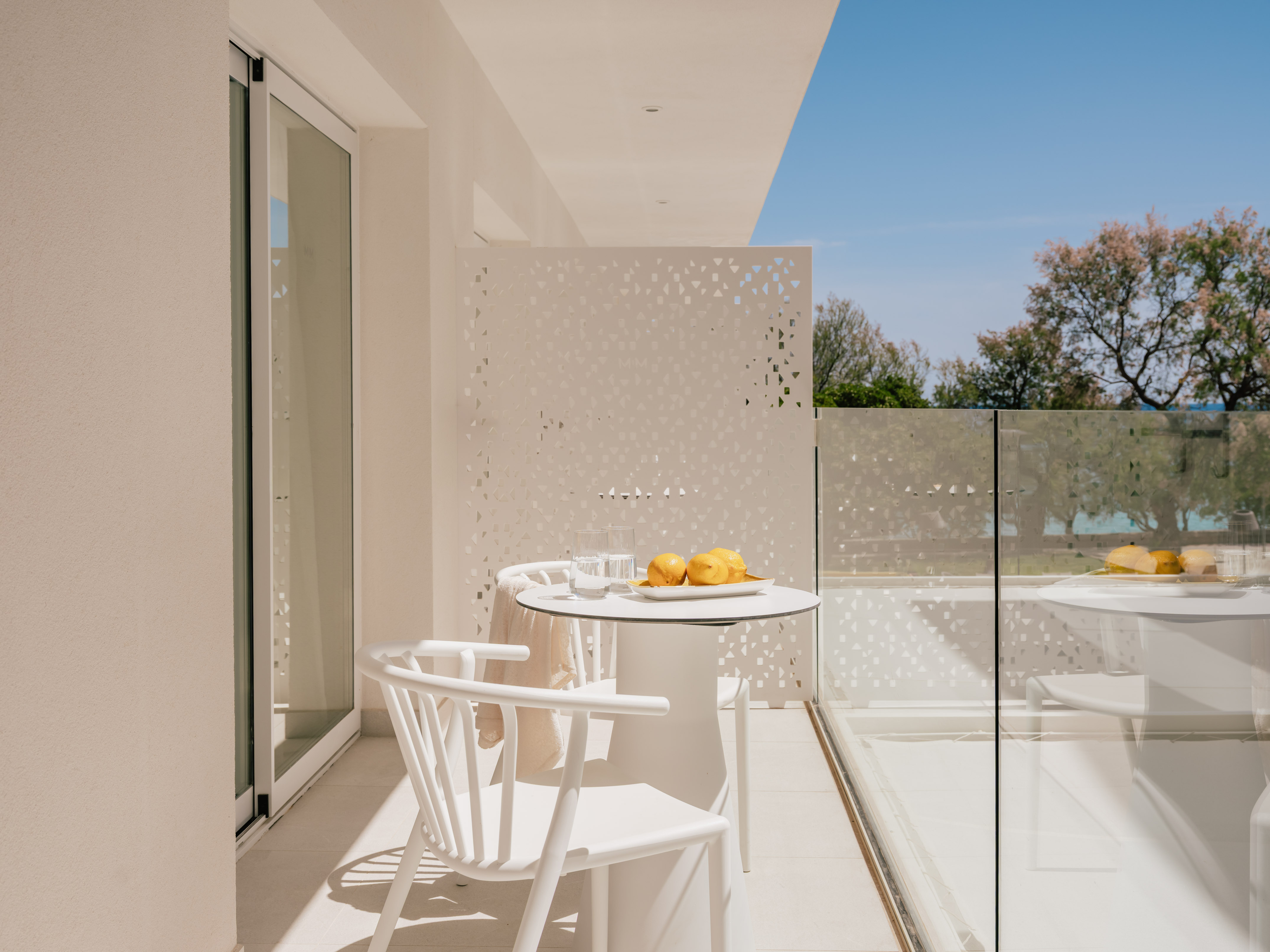 Modern white balcony with outdoor furniture, lemons, glass railing, and green trees.