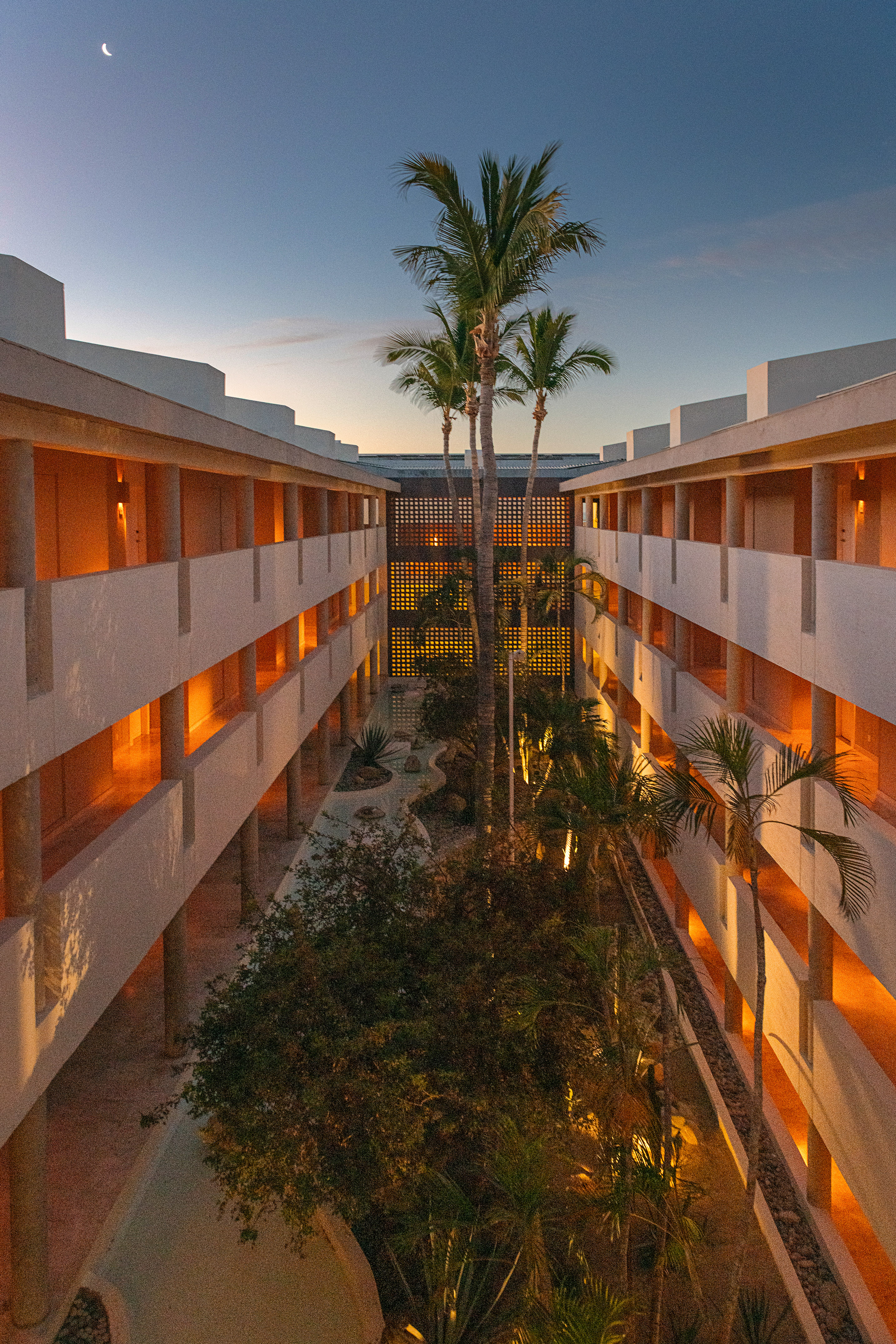 Modern hotel courtyard at dusk, illuminated rooms, palm trees, crescent moon.